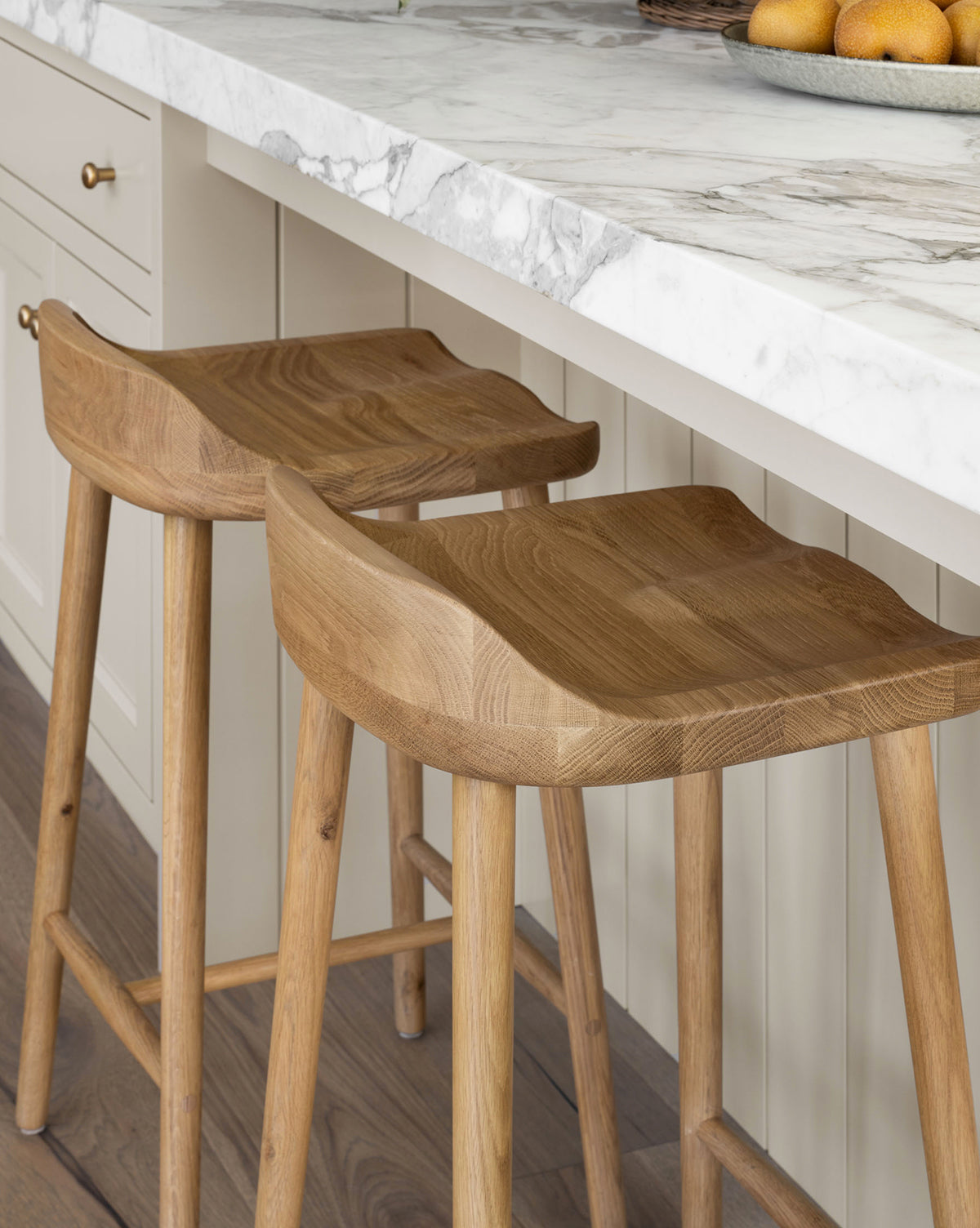 Two Makerspalm Portland Counter Stools with curved oak seats are set at a marble countertop in a modern kitchen with light cabinets and wood floors, McGee & Co.
