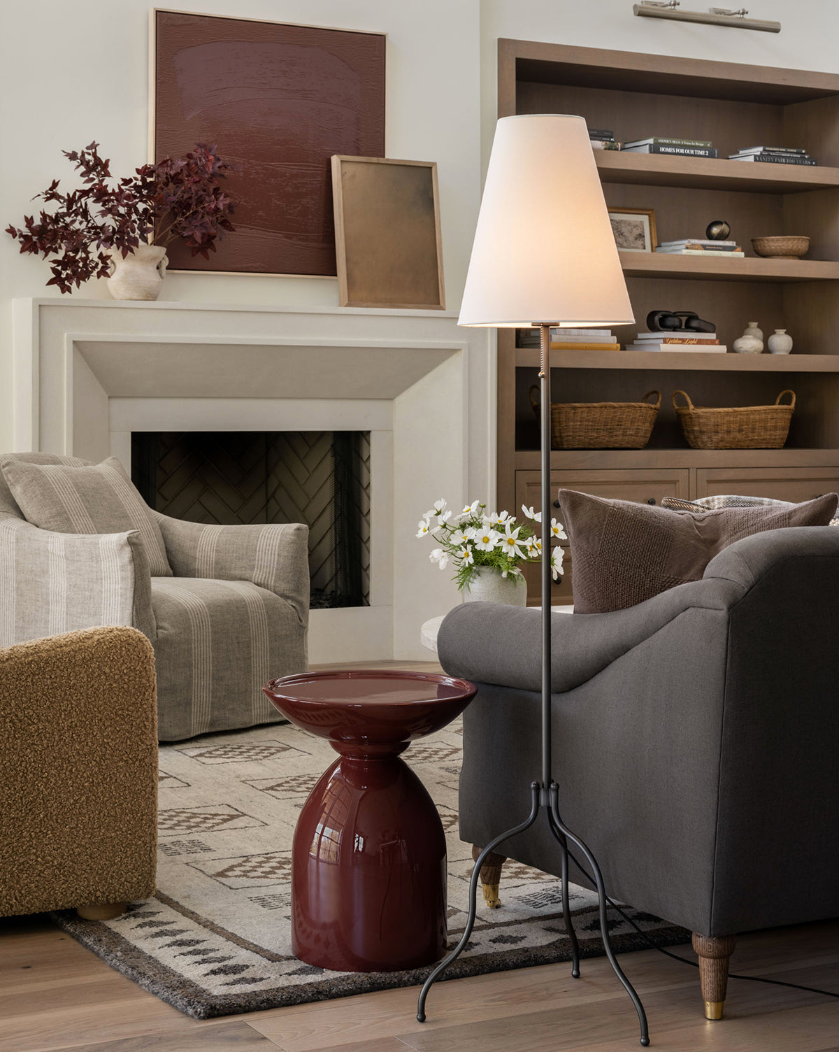 A cozy living room with a gray armchair, red side table, shelves of books and decor, and potted flowers by the armchair. The Windham Floor Lamp with a matte black base and white linen shade stands beside the fireplace, by McGee & Co.