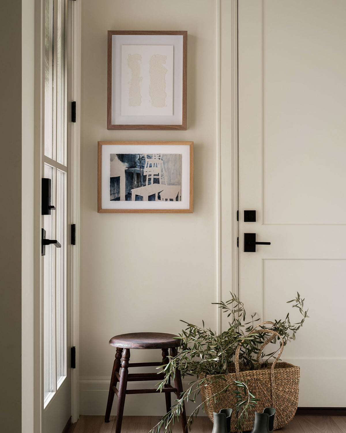 A small entryway with a white glass-paneled door, two framed fine art paper artworks, a wooden stool, and a woven basket of branches and greenery. Natural light brightens this tranquil Space Study.
