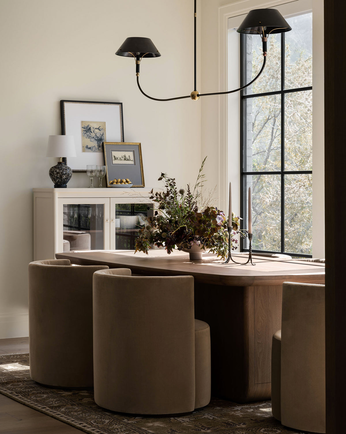 A modern dining room features a wooden table, Georgina Dining Chairs with barrel backs in velvet, a floral centerpiece, two black candle holders, large window, sideboard with framed art—all in neutral tones and natural light by McGee & Co.