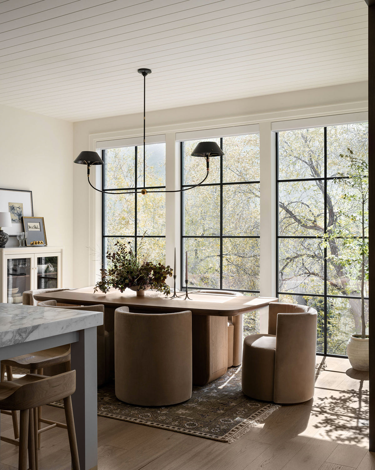 A sunlit dining area with large windows, a wooden table, round tan chairs, and a modern black chandelier highlights the Wayland Hand-Knotted Wool Rug, by McGee & Co. Decorative plants add charm beside a kitchen island with stools and a marble countertop.