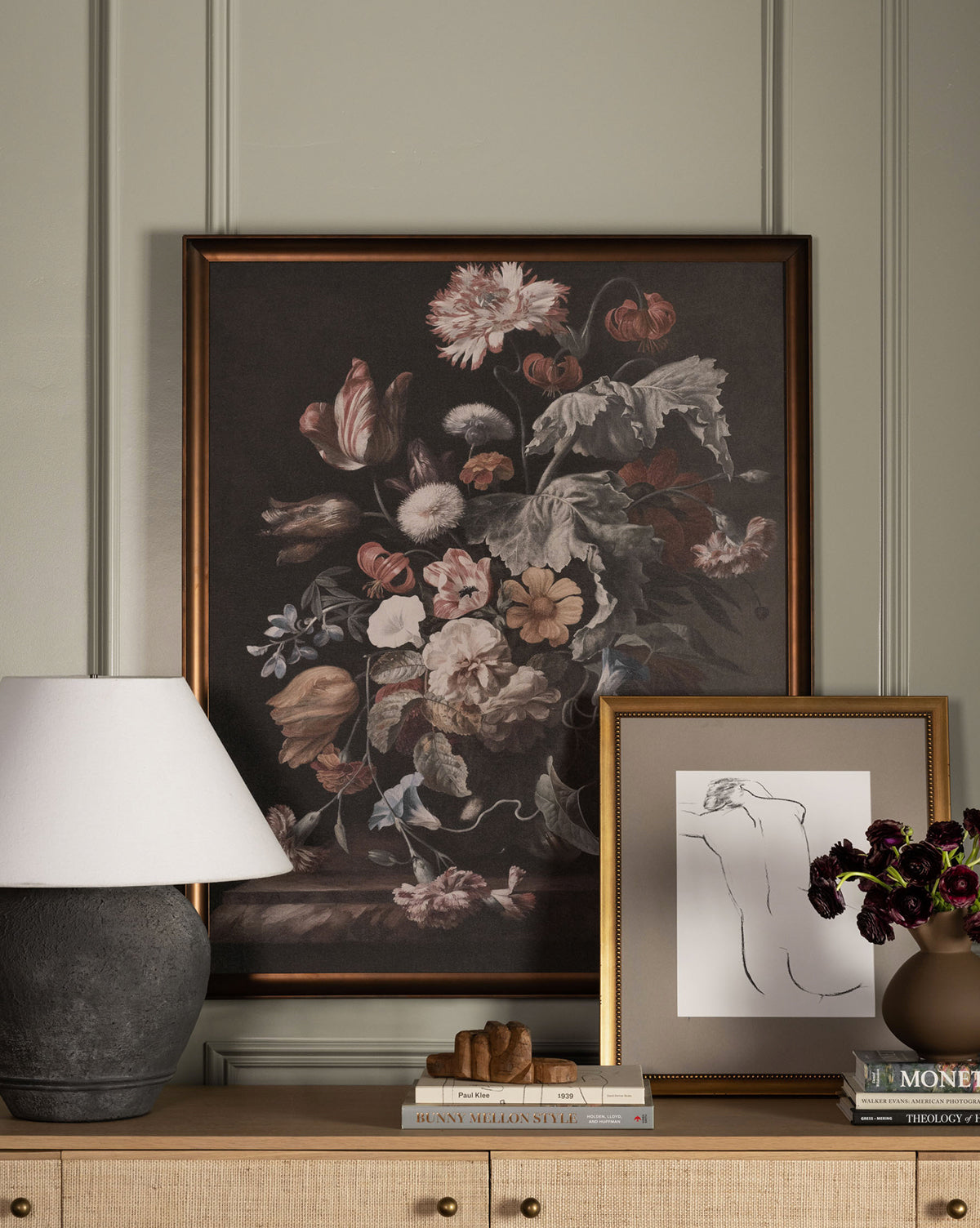 A wooden sideboard displays a table lamp, framed line drawing, stacked books, small sculpture, and flower vase; above hangs the Botanical Renaissance artwork.