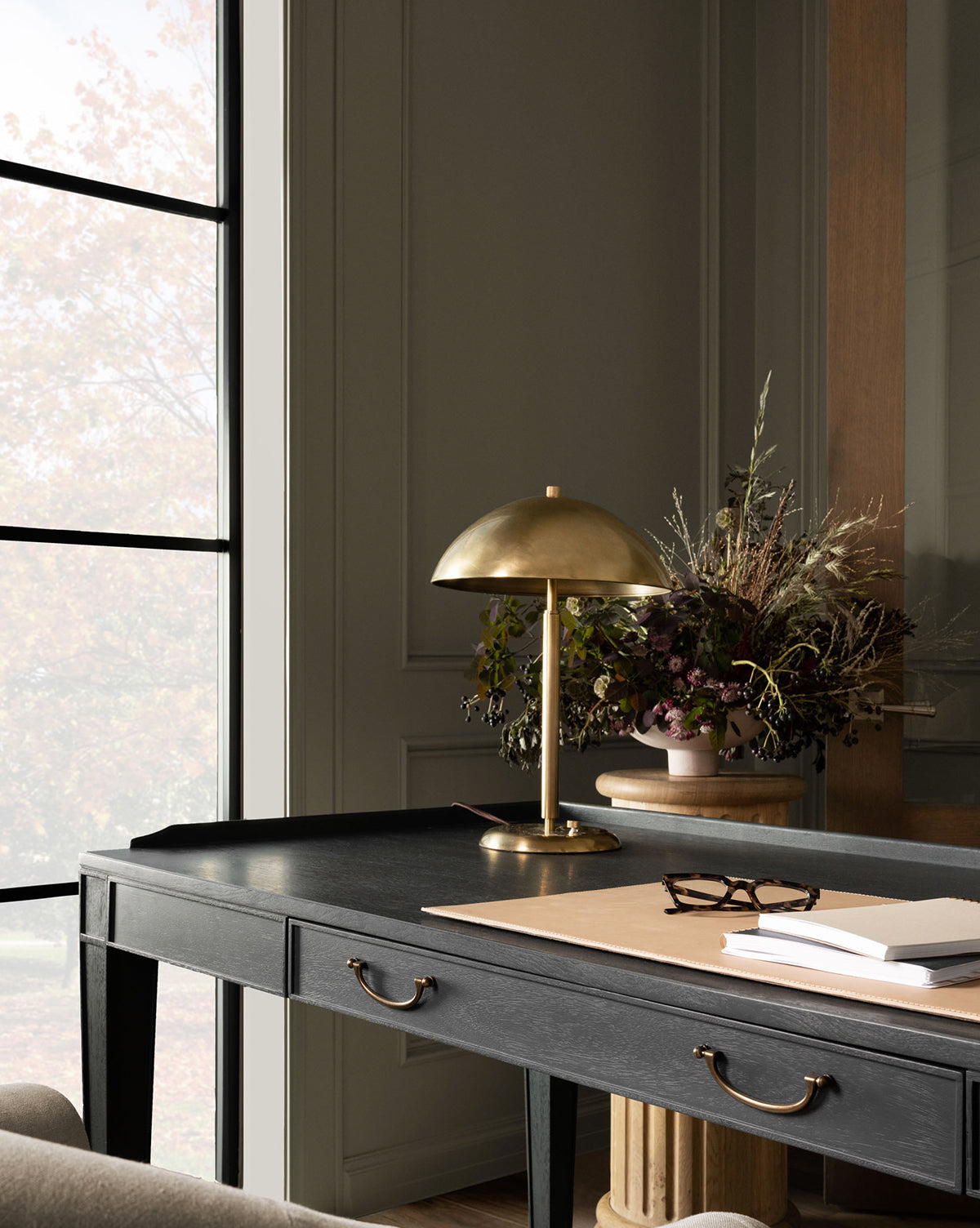 A black desk with brass handles holds the Aldorno Desk Table Lamp, a pair of glasses, a notebook, and papers. Behind the desk is a tall window and a pedestal with a vase of dried flowers in a modern, elegant room by McGee & Co.