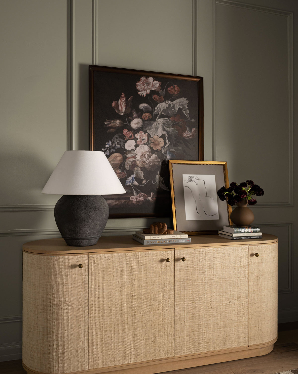 The Linu Woven Sideboard displays a gray lamp with a white shade, stacked books, a sculptural piece, a vase of dark flowers, and framed art featuring a floral painting and a minimalist line drawing by McGee & Co.