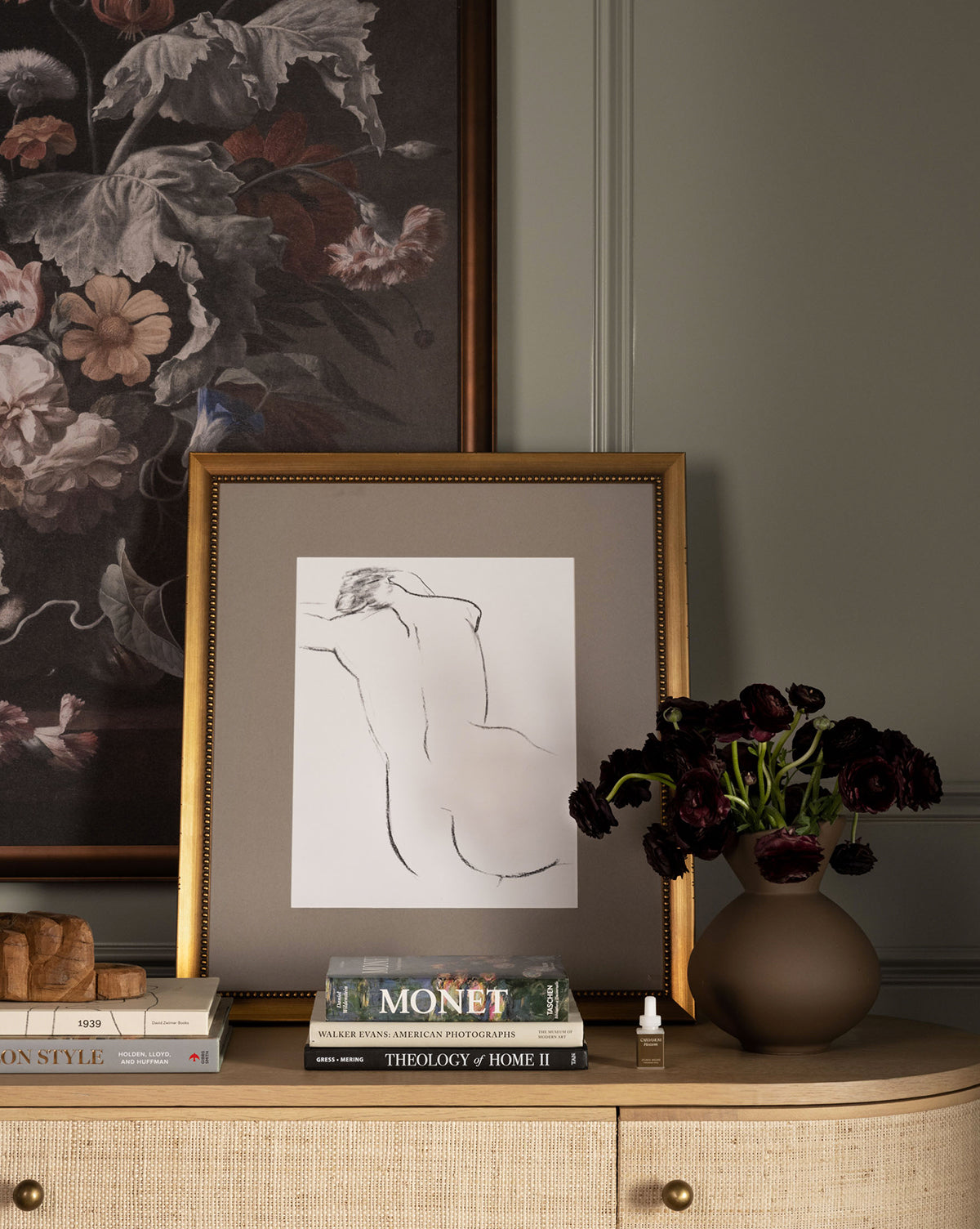 ‘Bare’, a minimalistic outline art nude in an antique gold frame, sits on a beige sideboard beside books and a brown vase with dark flowers, while a floral painting hangs on the wall behind.