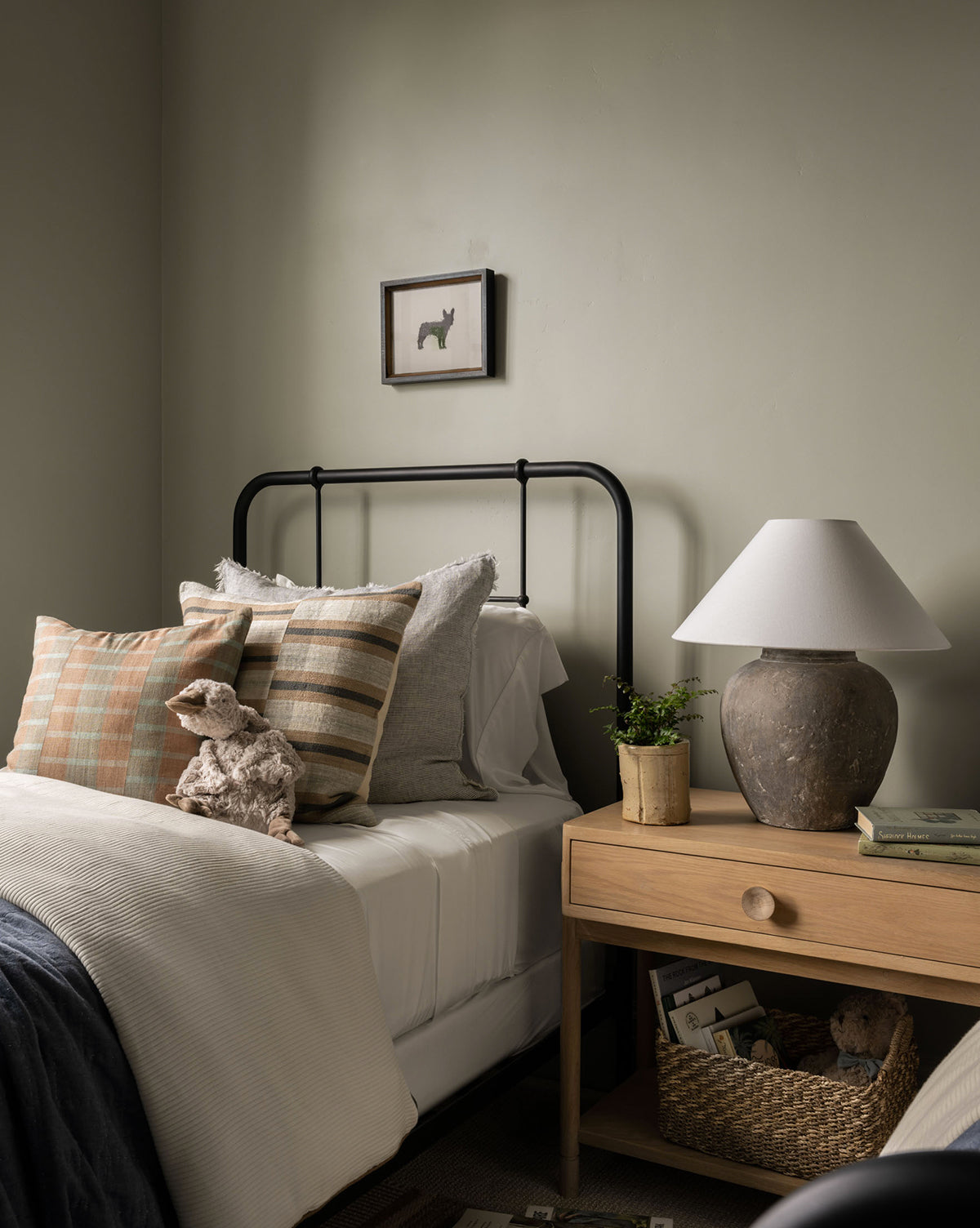 A cozy bedroom features a metal-frame bed with neutral bedding, plaid pillows, a stuffed sheep, wooden nightstand with rustic lamp, small plant, books, basket below, and Scribbled Dog wall art displayed above the bed.