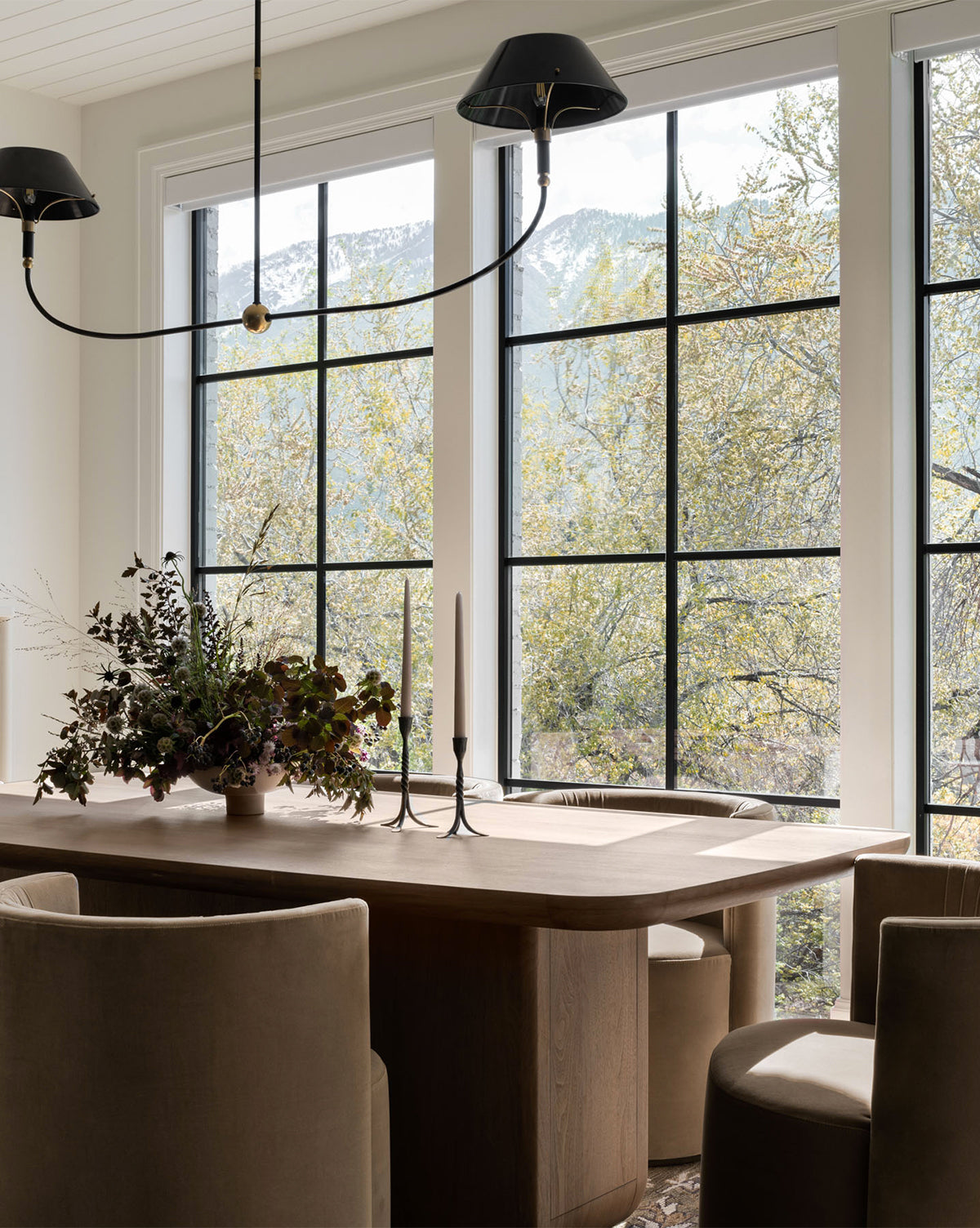 A modern dining room features the Cheswick Dining Table, beige chairs, decorative candlesticks, and a floral arrangement. Large windows reveal leafy trees and mountains, bathing the space in natural light by McGee & Co.