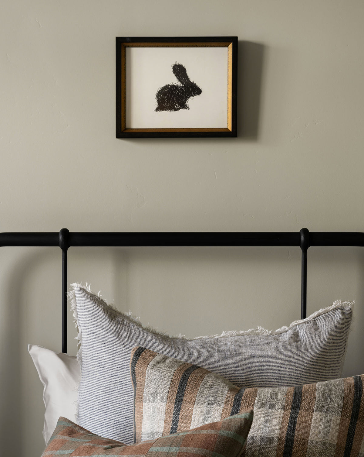 A framed Scribbled Rabbit hangs on a beige wall above a black metal bed. The bed features striped and plaid pillows in gray, beige, and brown tones.