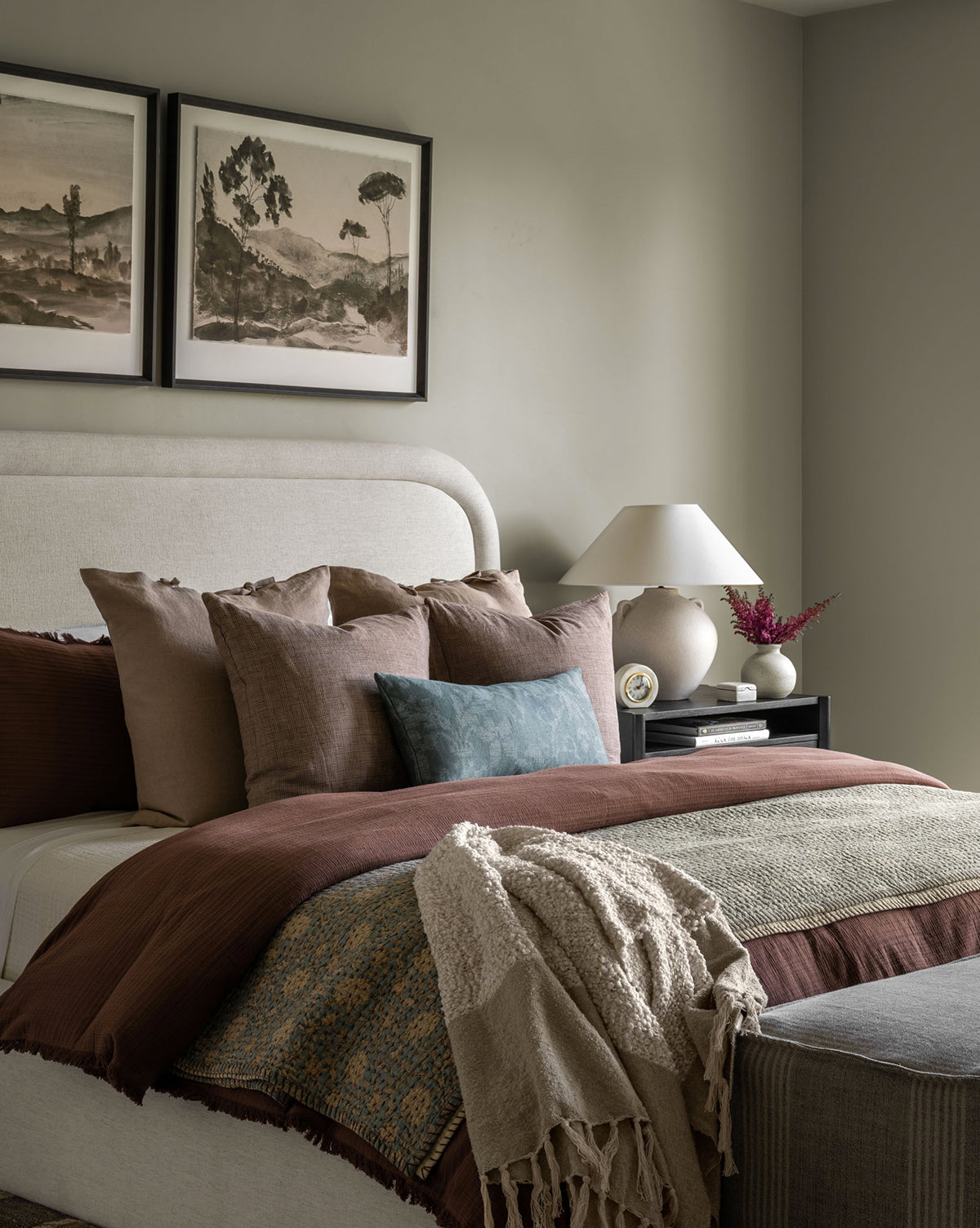 A cozy bedroom showcases a beige upholstered bed with layered pillows and the Hendricks Chocolate Cotton Duvet Cover in brown, blue, and cream. A nightstand displays a lamp, books, and flowers, with two landscape art prints above the bed.