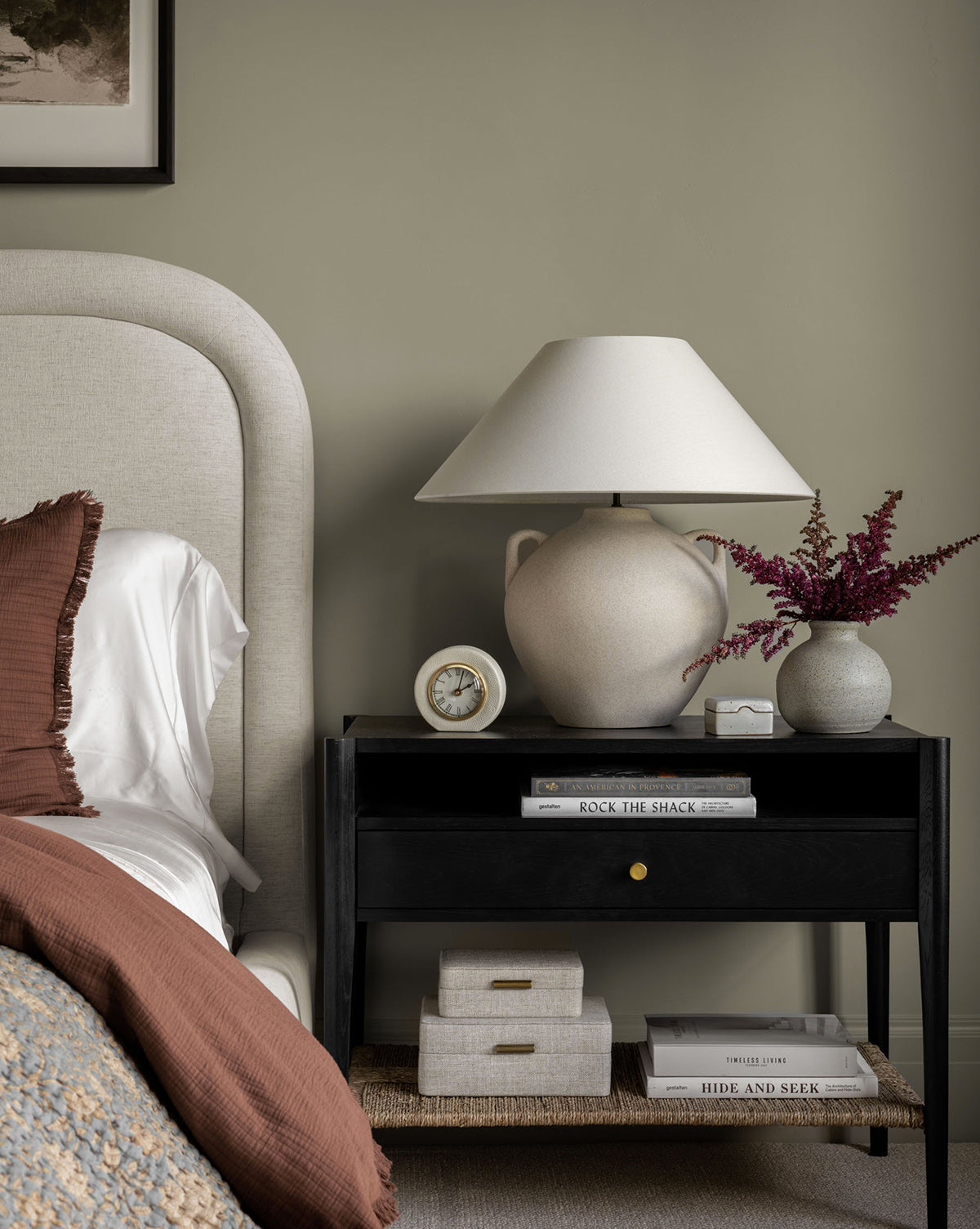 A cozy bedroom features a beige upholstered bed, white bedding, and a brown pillow. A black nightstand holds books, a large lamp, a small vase with purple flowers, and the Shagreen Table Clock. Neutral decor adds to the calm atmosphere.
