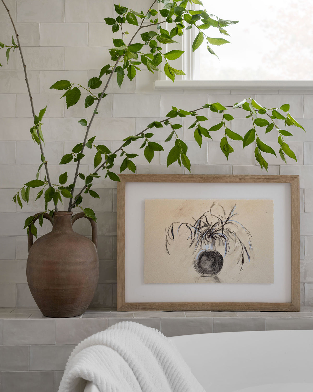Willow Branches arranged in a ceramic vase sit on a tiled ledge beside a framed botanical print; a white bathtub and rolled towel can be seen in the foreground.