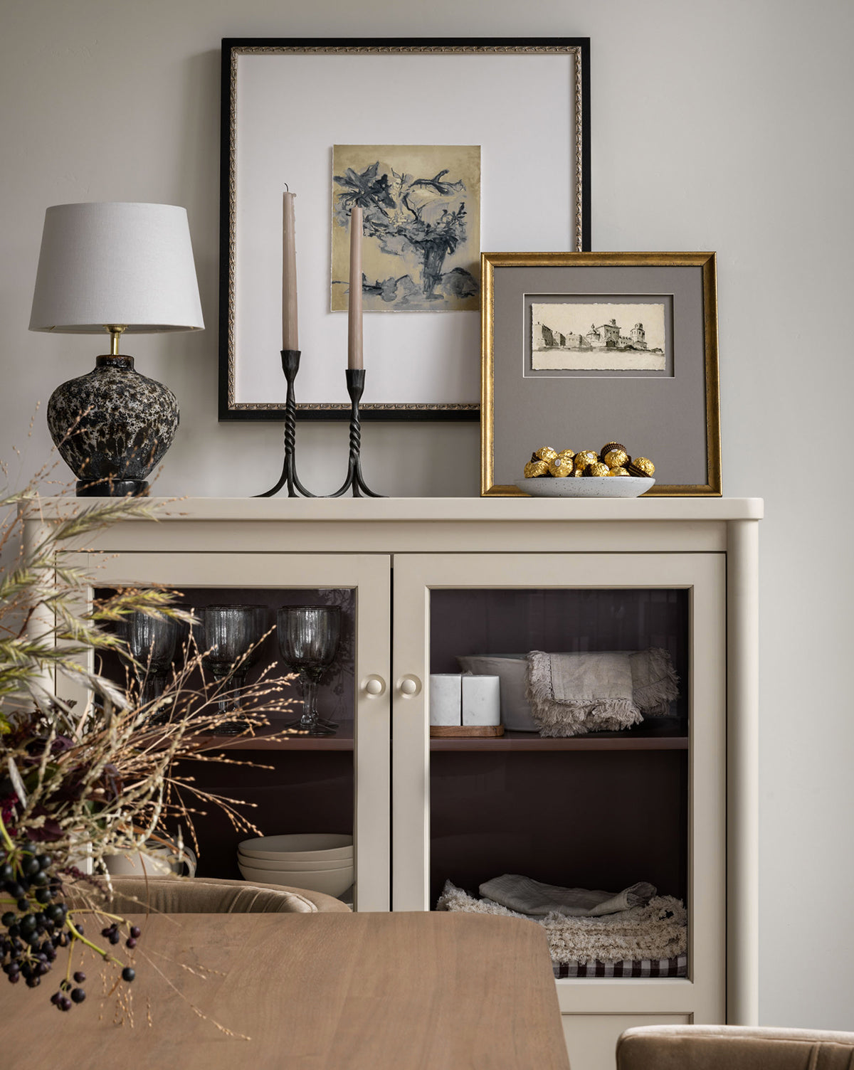 A cream-colored cabinet with glass doors holds folded linens and glassware. On top are a lamp, framed botanical art, a bowl of gold decorative balls, and Abstracted Votives. Dried flowers sit on a nearby wooden table.