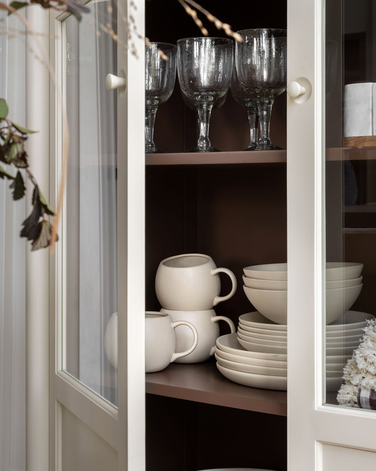 A cream-colored cabinet with glass doors displays stacked white ceramic cups and plates, including the Luana Bowl on the bottom shelf, and clear goblets above. The modern neutral scene features dried foliage to the left, by McGee & Co.