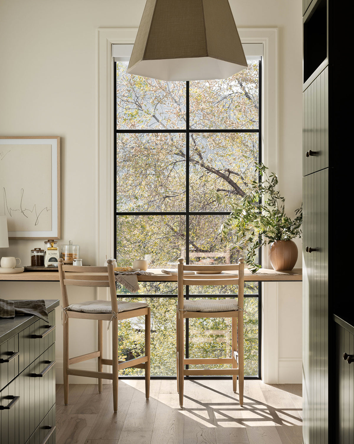 A bright kitchen nook features two Romany Counter Stools with linen cushions by a large window overlooking leafy trees. Natural light fills the space, while plants and simple decor create a cozy, inviting atmosphere by McGee & Co.