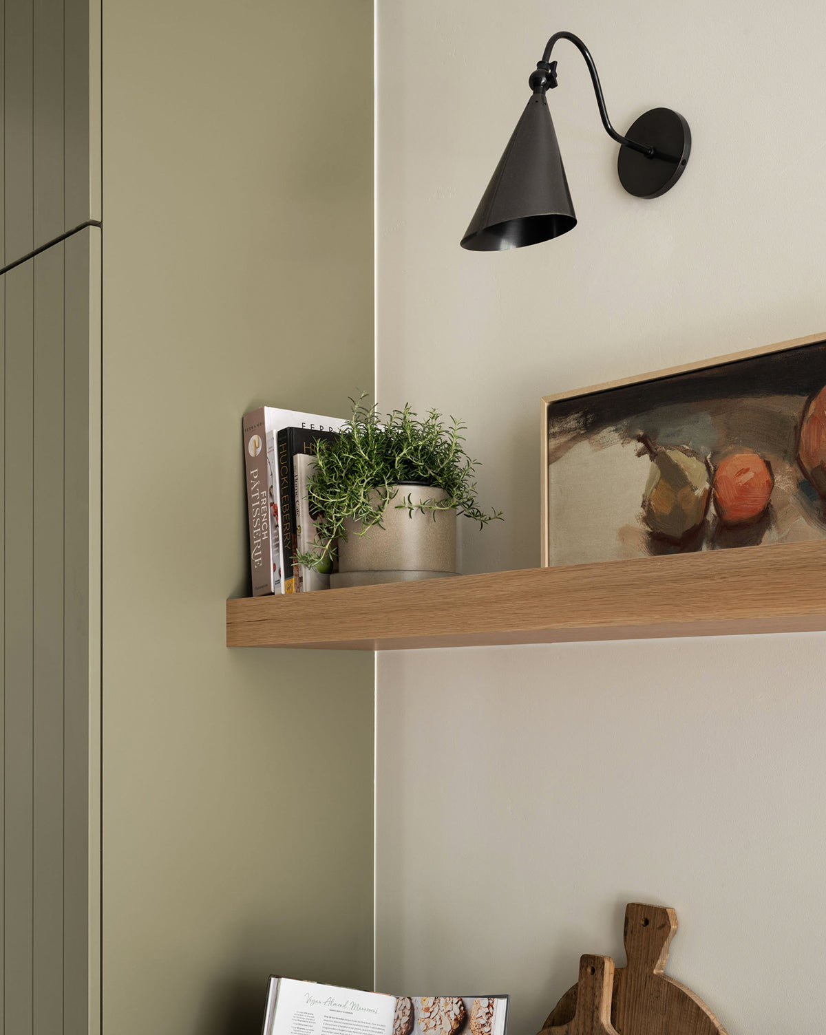 A wooden shelf displays cookbooks, a potted plant, and the Paired Pears wall art under a black sconce. Nearby, part of a cabinet and cutting boards finish this charming kitchen vignette.