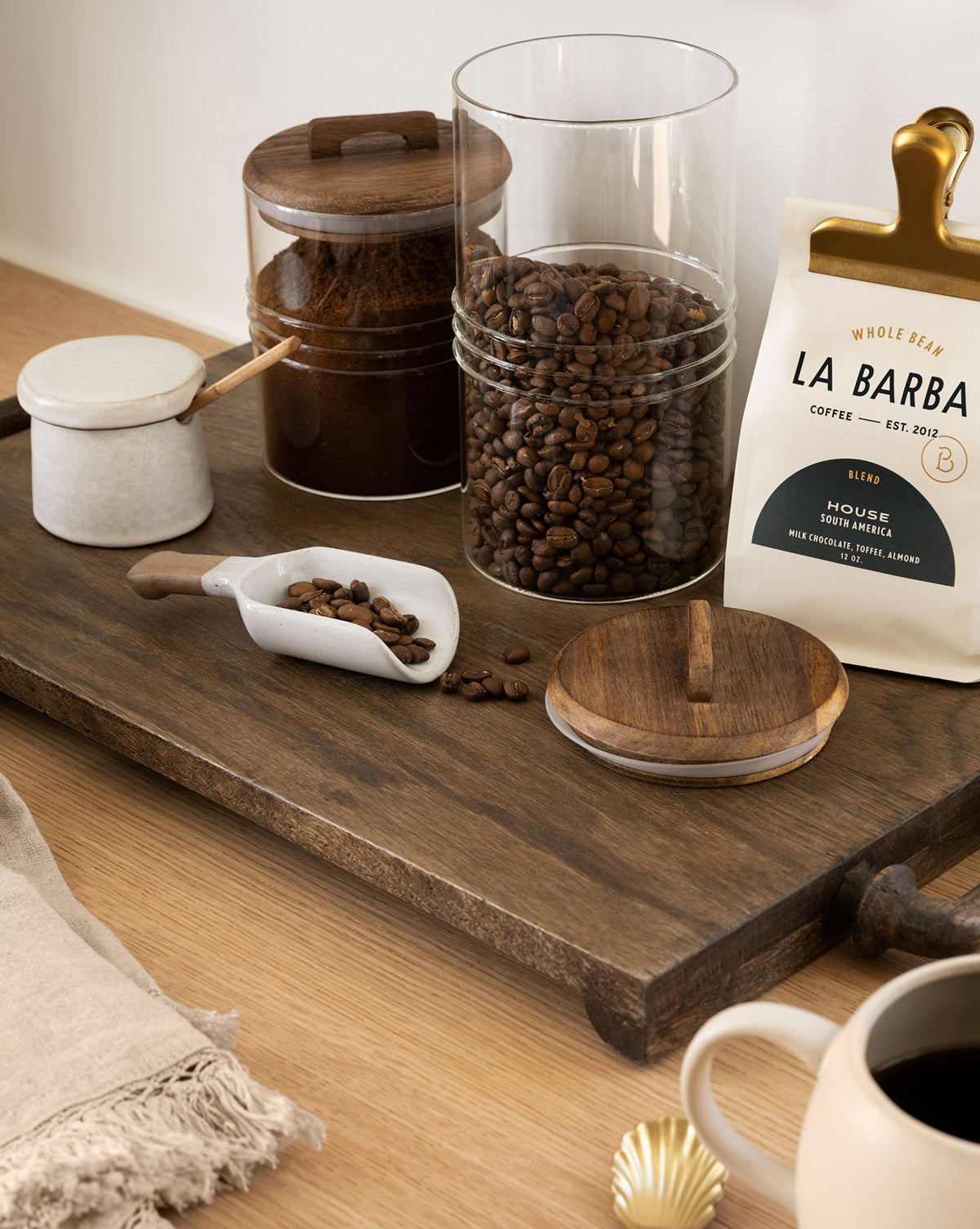 A wooden tray holds jars of coffee beans and ground coffee, a coffee scoop, a bag labeled La Barba Coffee, and the Malco Salt Cellar. Nearby on the light wood table are a cup of black coffee and a gold spoon.