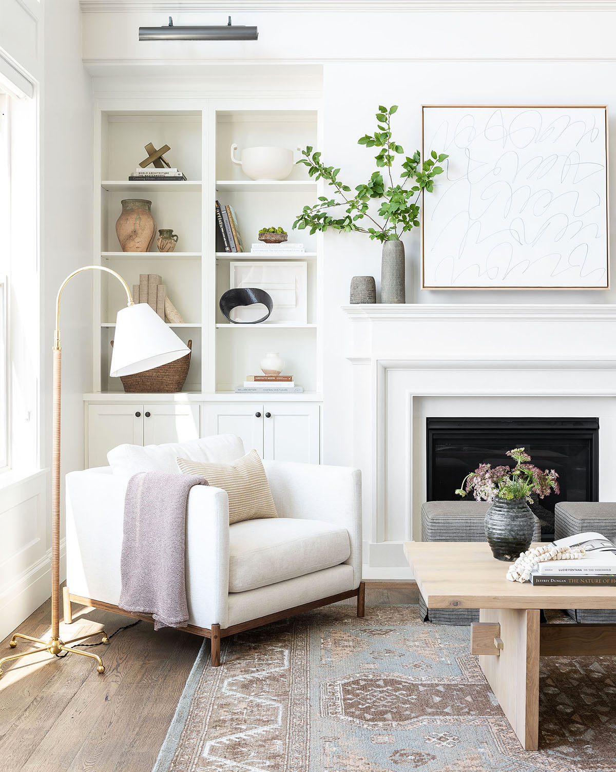 Bright, modern living room featuring a white armchair, light throw blanket, floor lamp, wood coffee table, built-in shelves with decor, and a fireplace topped with the Scattered Thoughts artwork in a natural floater frame and a vase of green branches.