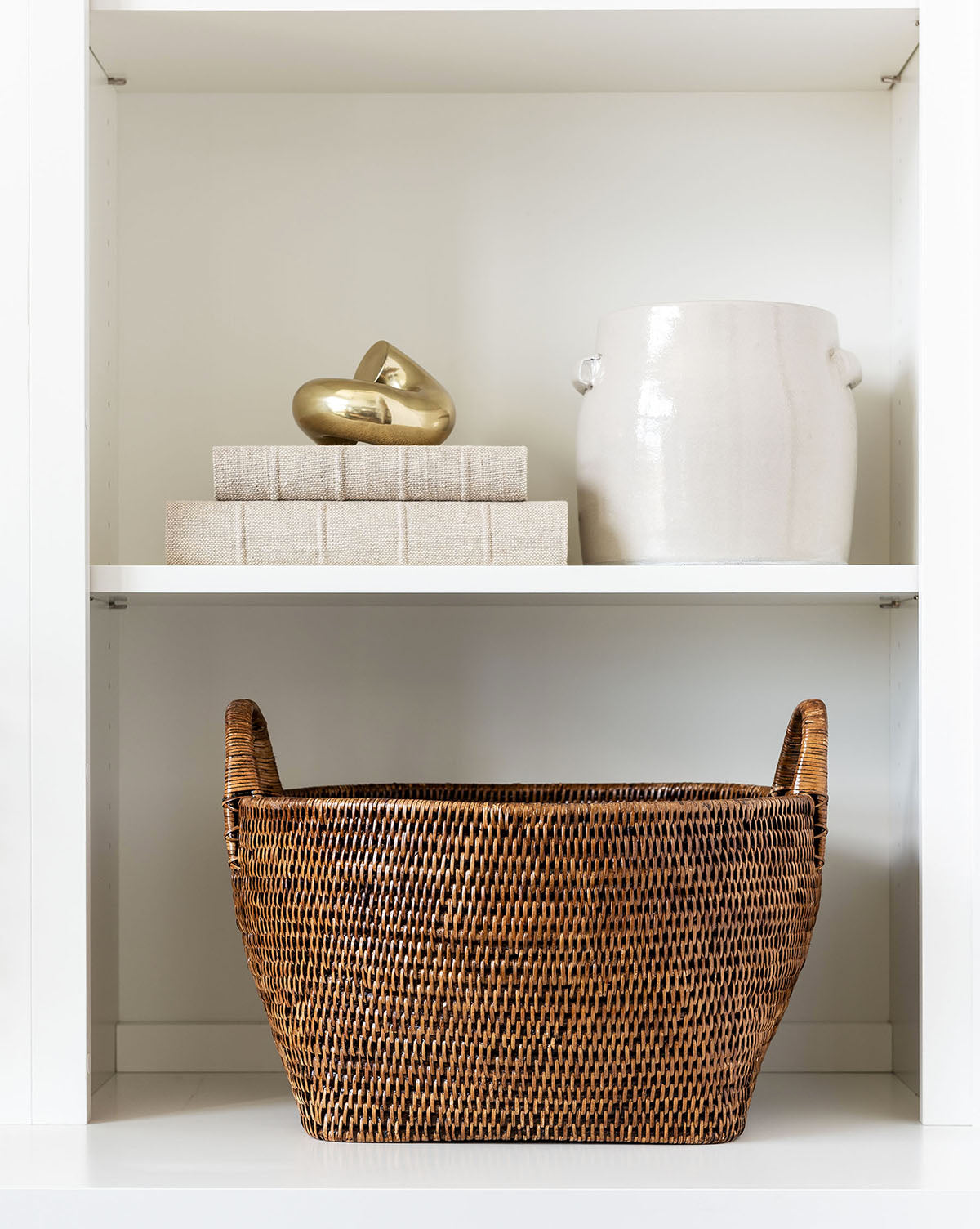 A shelf displays a white vase, three Handcrafted Linen Books stacked together, a gold abstract sculpture, and a large woven basket with handles on the lower shelf.
