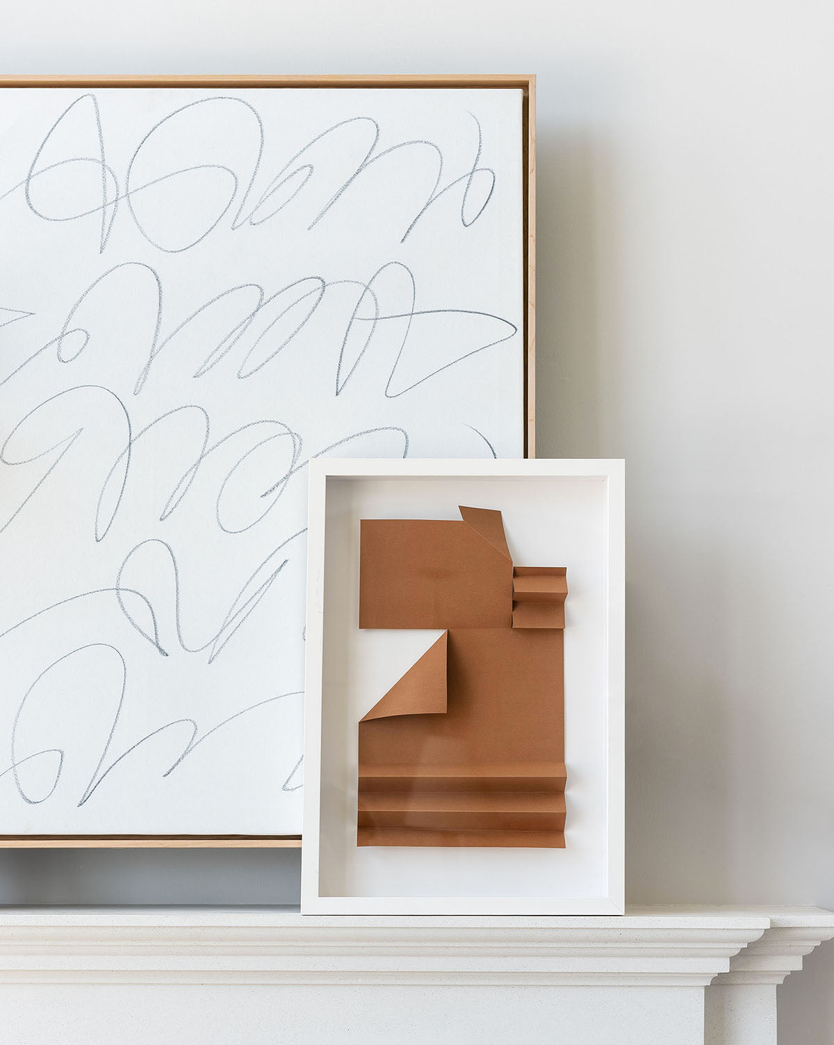 The Scattered Thoughts framed abstract digitally printed canvas featuring gray scribbles leans against a wall behind a smaller frame with folded brown paper, both pieces of wall art displayed on a white mantelpiece.