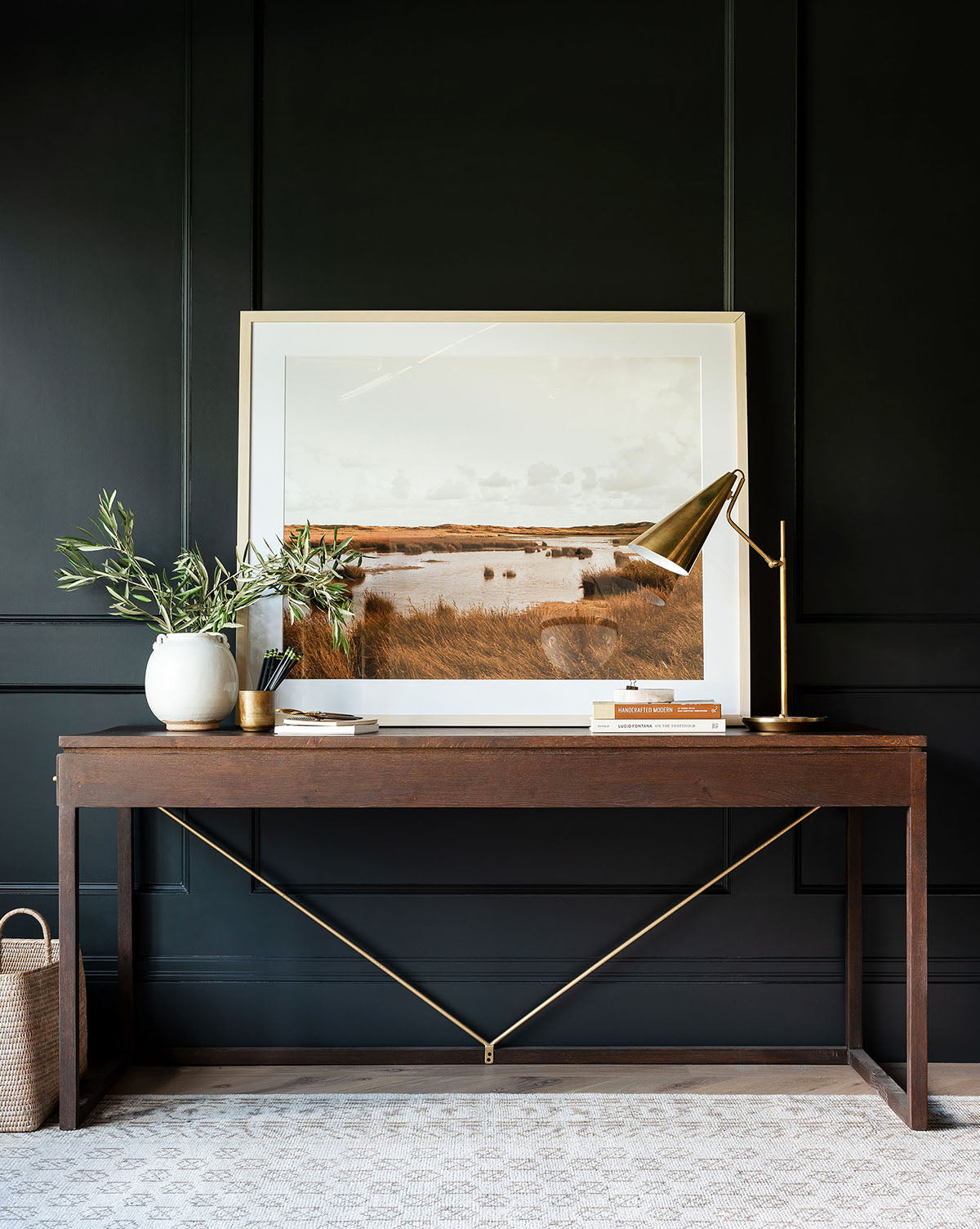 A dark wood console table with gold accents holds a plant, books, a gold lamp, and a framed Pond & Meadow print against a black paneled wall. A basket rests on the floor nearby.