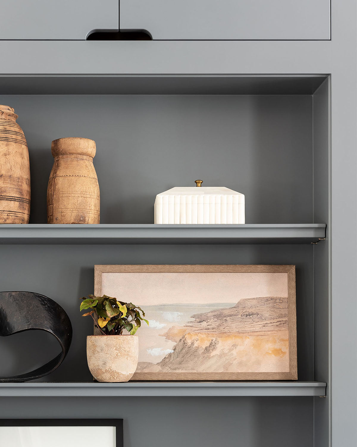 A gray shelving unit showcases designer decor: two wooden vases, a Plateau Lid Box, a black sculpture, a small potted plant, and a framed landscape painting of cliffs and water.
