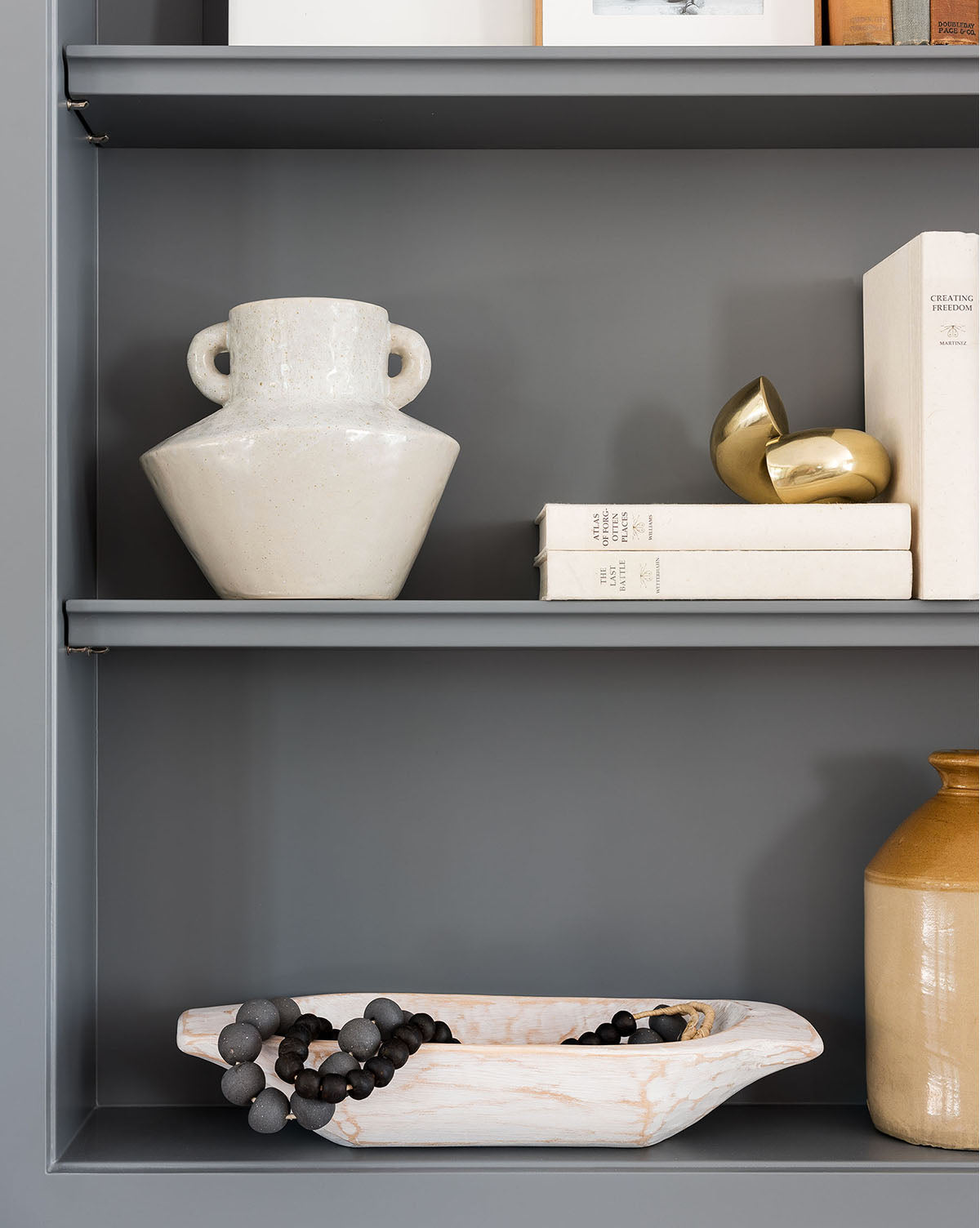A gray shelf displays the cream ceramic Malaga Vase, stacked white books, two gold decorative spheres, a beige and brown streaked bowl with a black beaded necklace, and a tan glazed vase.