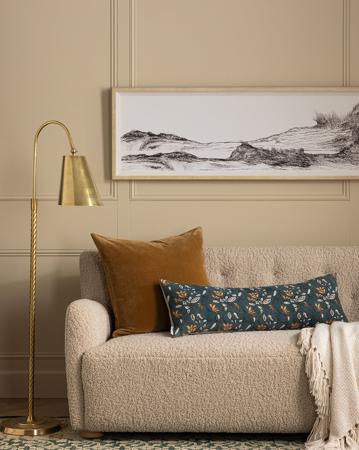 A cozy beige textured sofa with a gold pillow and a long blue floral cushion sits beside a brass floor lamp. At the Seashore artwork hangs on the beige paneled wall, and a cream throw blanket is draped over the sofa arm.
