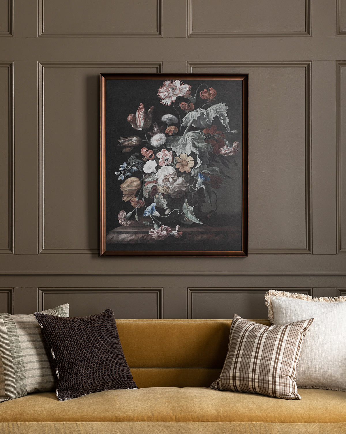 Botanical Renaissance, a framed floral still life painting, is displayed on a taupe paneled wall above a mustard-yellow sofa adorned with assorted patterned and textured throw pillows.