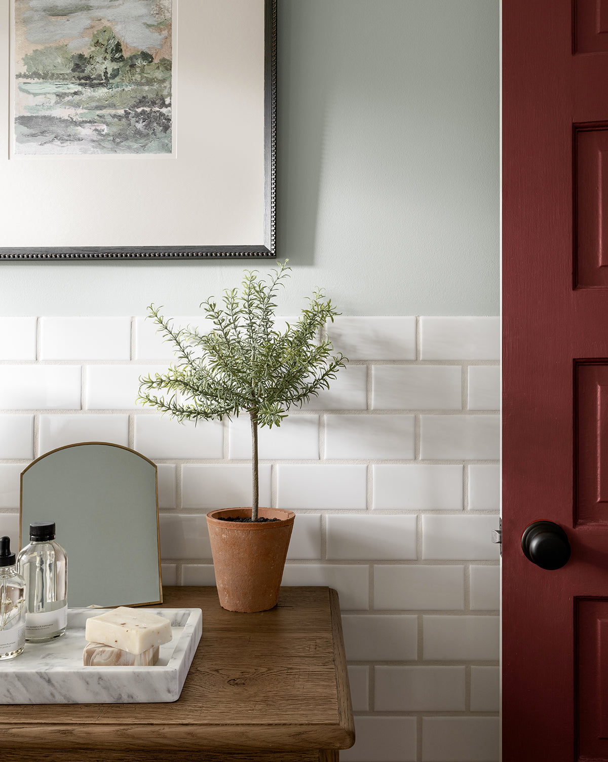 A small potted plant sits on a wooden table with toiletries, beside white subway tile walls. Above is Forest Meadow, a framed fine art print on textured paper; to the right is a red door with a black handle.