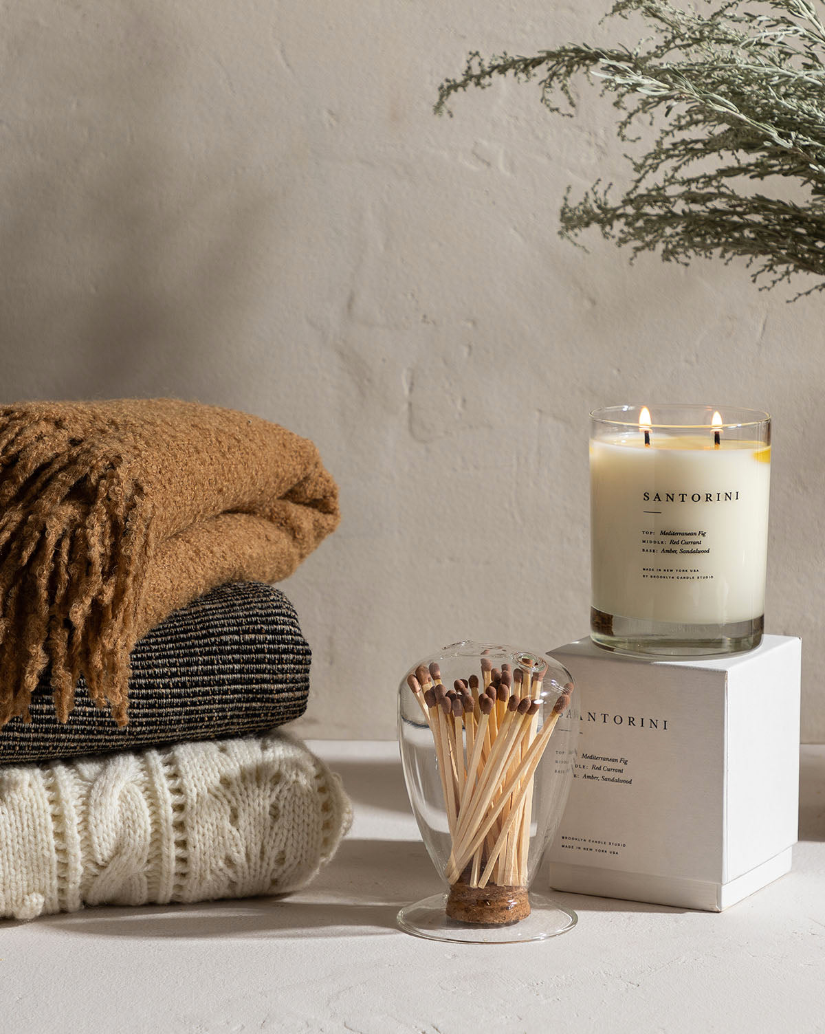 A cozy scene features a stack of folded sweaters, a tan scarf, a glass match holder, and the soy wax Santorini Candle on a white box, filling the air with Mediterranean fig notes against a neutral wall.