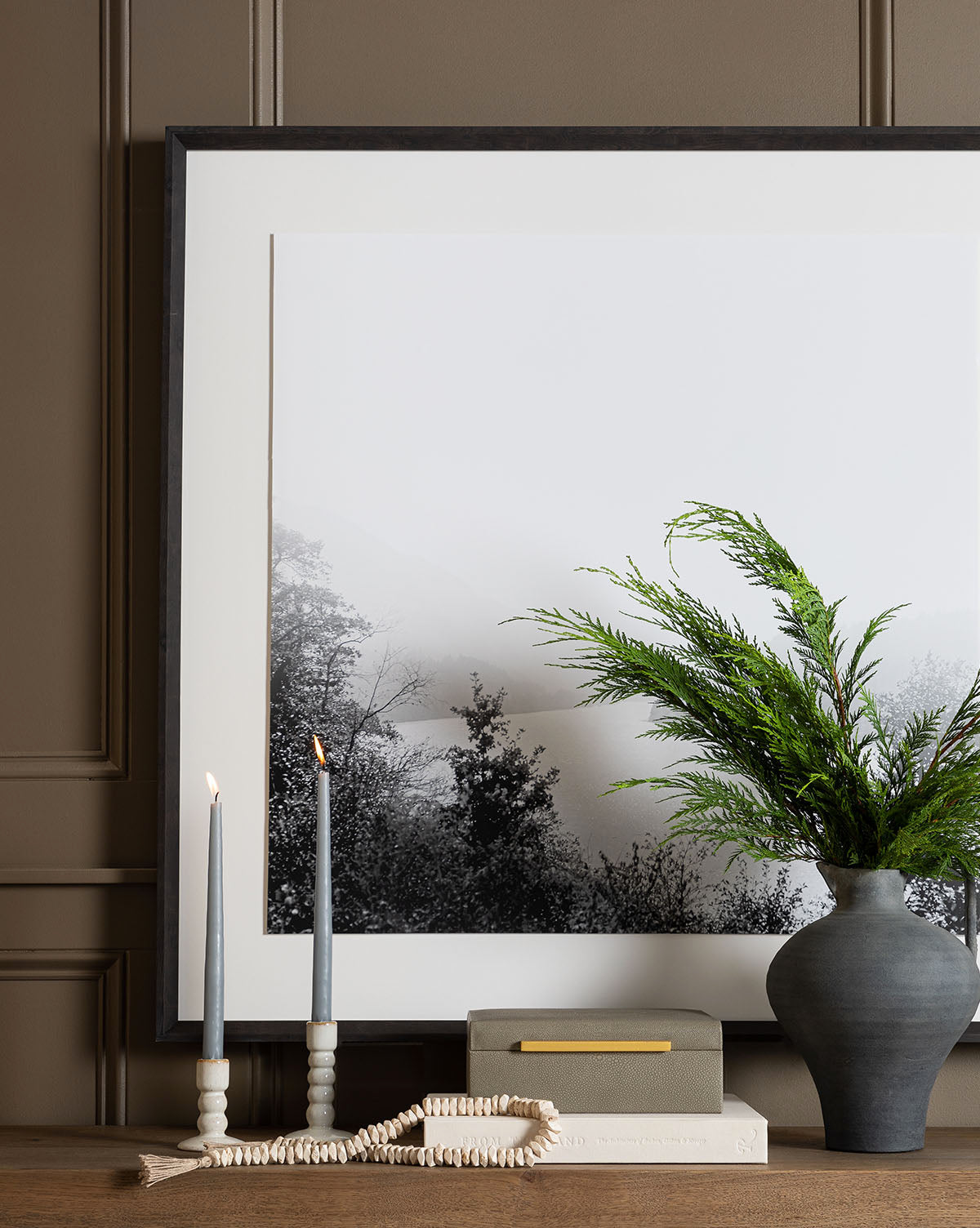 Framed black-and-white landscape art sits behind a vase with green foliage, warm candlelight from Light Gray Taper Candles (Set of 12) in holders, books, and a decorative bead garland on a wood surface against a brown paneled wall.