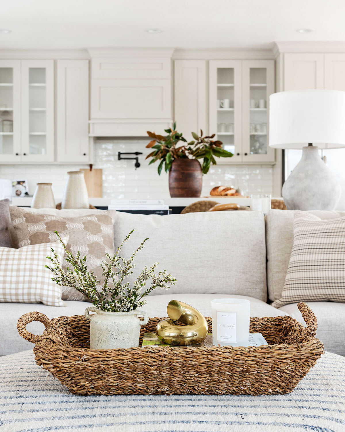 A woven tray with a potted plant, gold decor, and a candle sits on a beige ottoman before a neutral sofa with plaid pillows. In the bright white kitchen behind, glass cabinets flank a Dual Handled Vase for added texture.