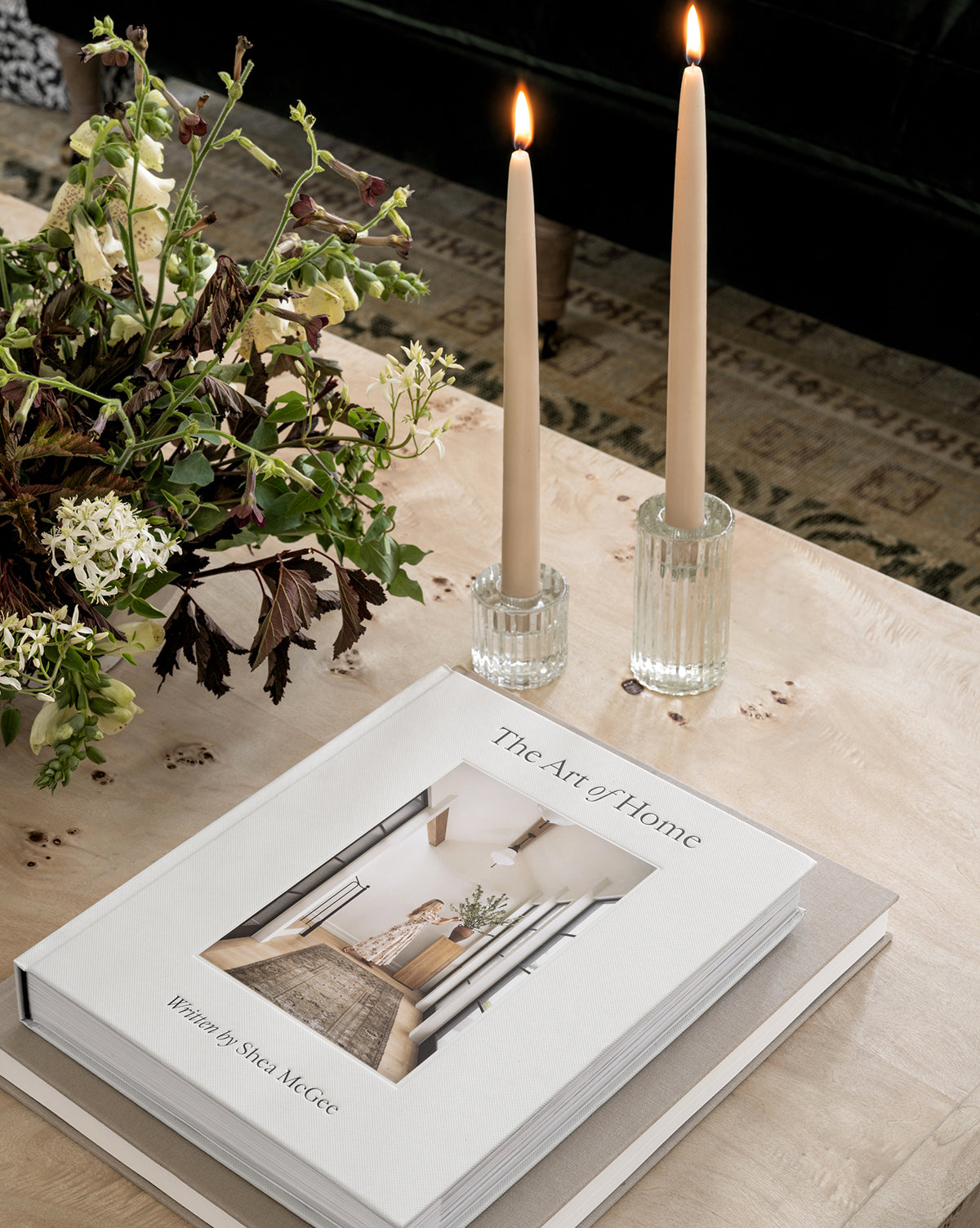 A coffee table features The Art of Home by Shea McGee, a floral bouquet, and two lit beige candles—elegant accents for those passionate about home decor and creating an inviting atmosphere.