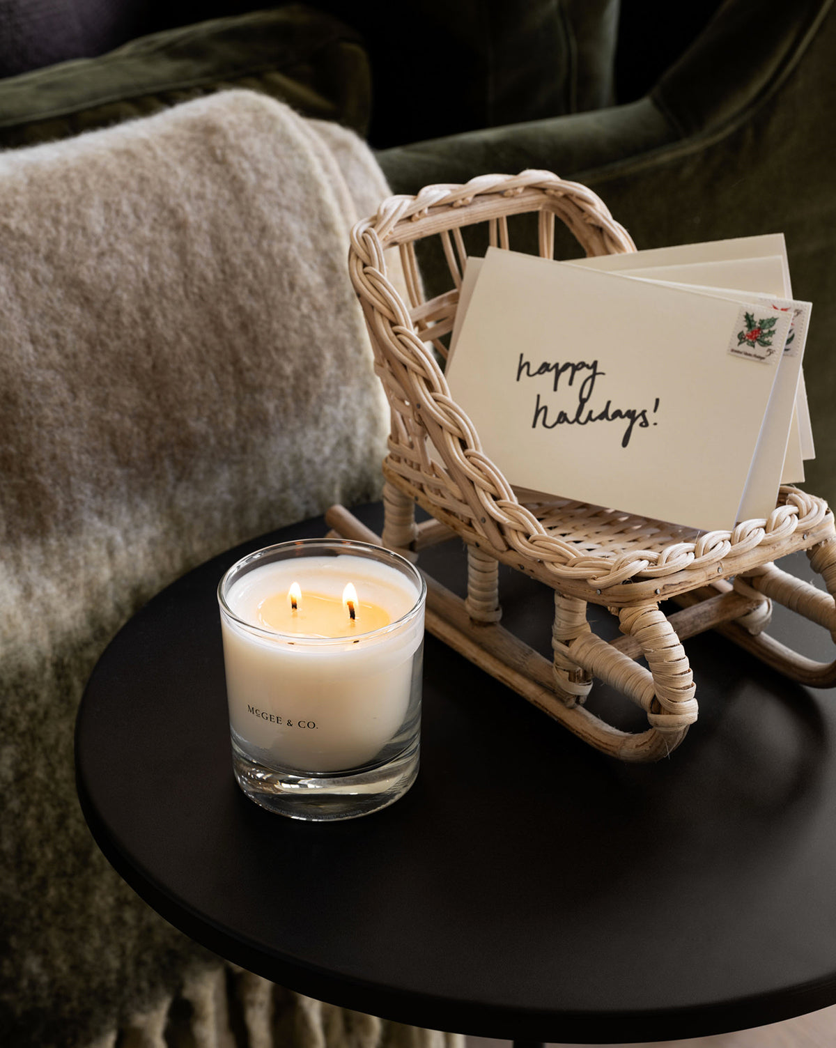 A glass candle with two flames sits on a black table beside the Woven Rattan Sleigh in a natural finish, holding holiday cards—one says “happy holidays!” A textured blanket and green cushion complete the festive decor by McGee & Co.