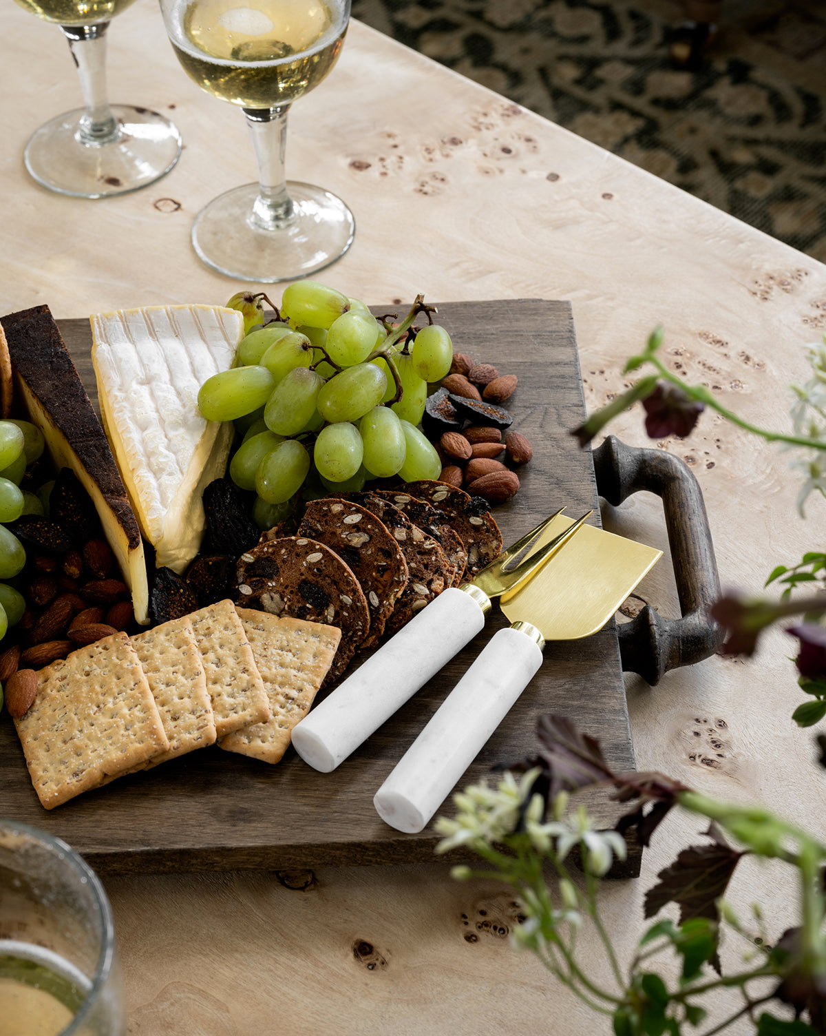 The Fordham Board, crafted from solid oak, beautifully presents cheese, green grapes, crackers, nuts, and seeded crisps. A marble-handled cheese knife and fork accompany it, with two glasses of white wine placed nearby by McGee & Co.