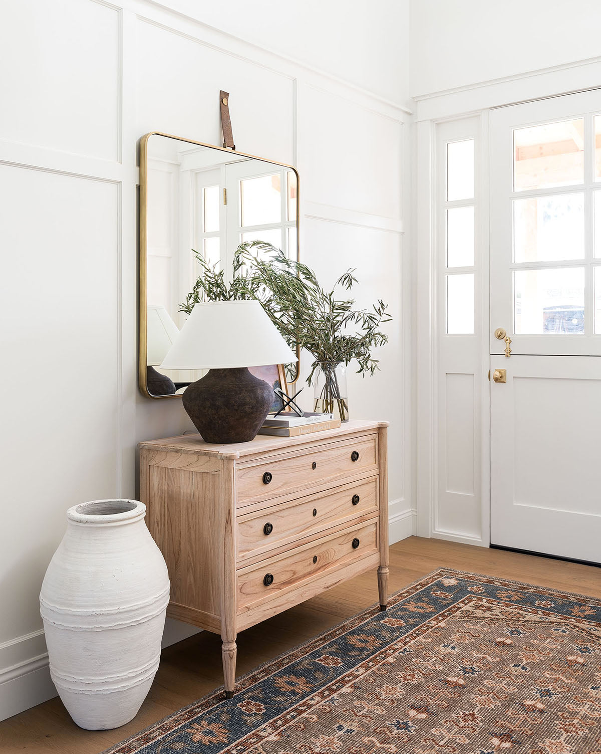 A light wood dresser with brass hardware, a brown lamp, a leafy plant in a vase, and the Laila Rectangle Mirror above sits near a white front door. A tall white ceramic vase and patterned rug complete the stylish entryway by McGee & Co.