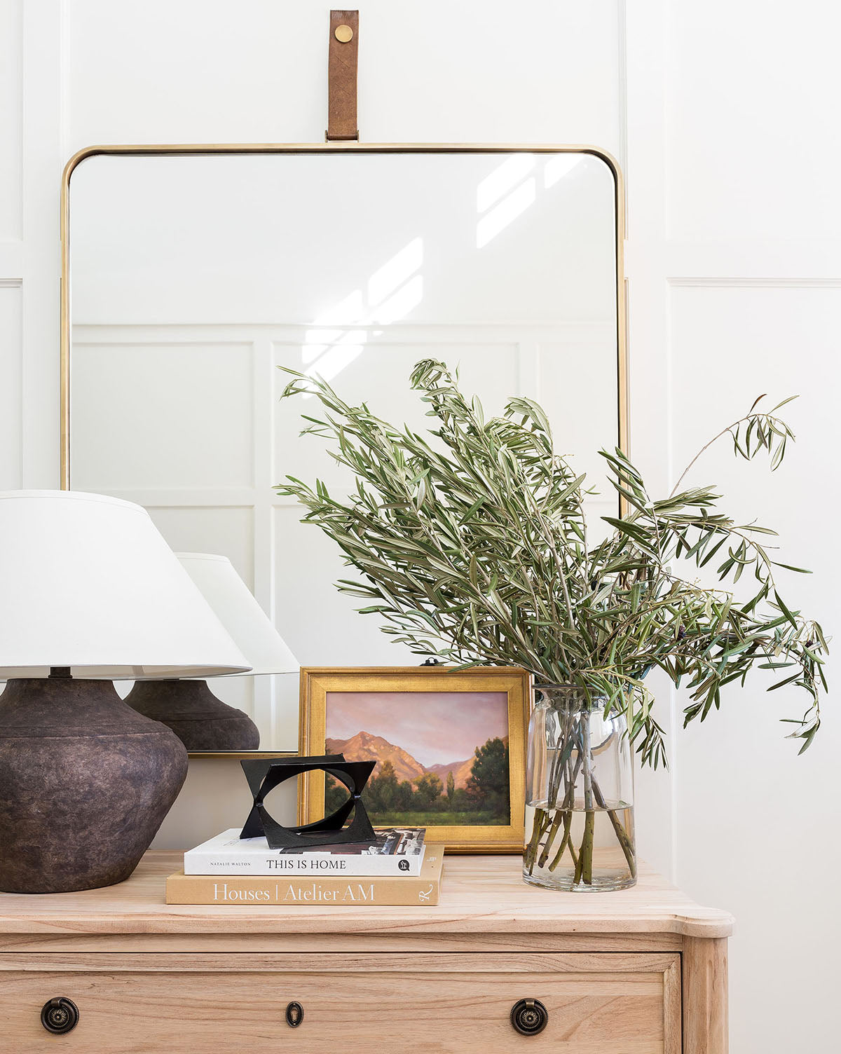 A wooden dresser with brass hardware is styled with a dark lamp, glass vase of leafy branches, small framed landscape painting, decor accents, and two stacked books. The Laila Rectangle Mirror is mounted on the wall behind in a gold frame by McGee & Co.