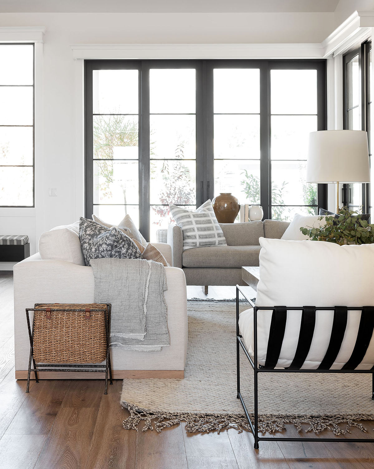 Bright, modern living room with large black-framed windows, a Vernon Sofa 92 with throw pillows and blanket, woven basket, light rug on wooden floors, alder wood accents, and a floor lamp beside a potted plant by McGee & Co.