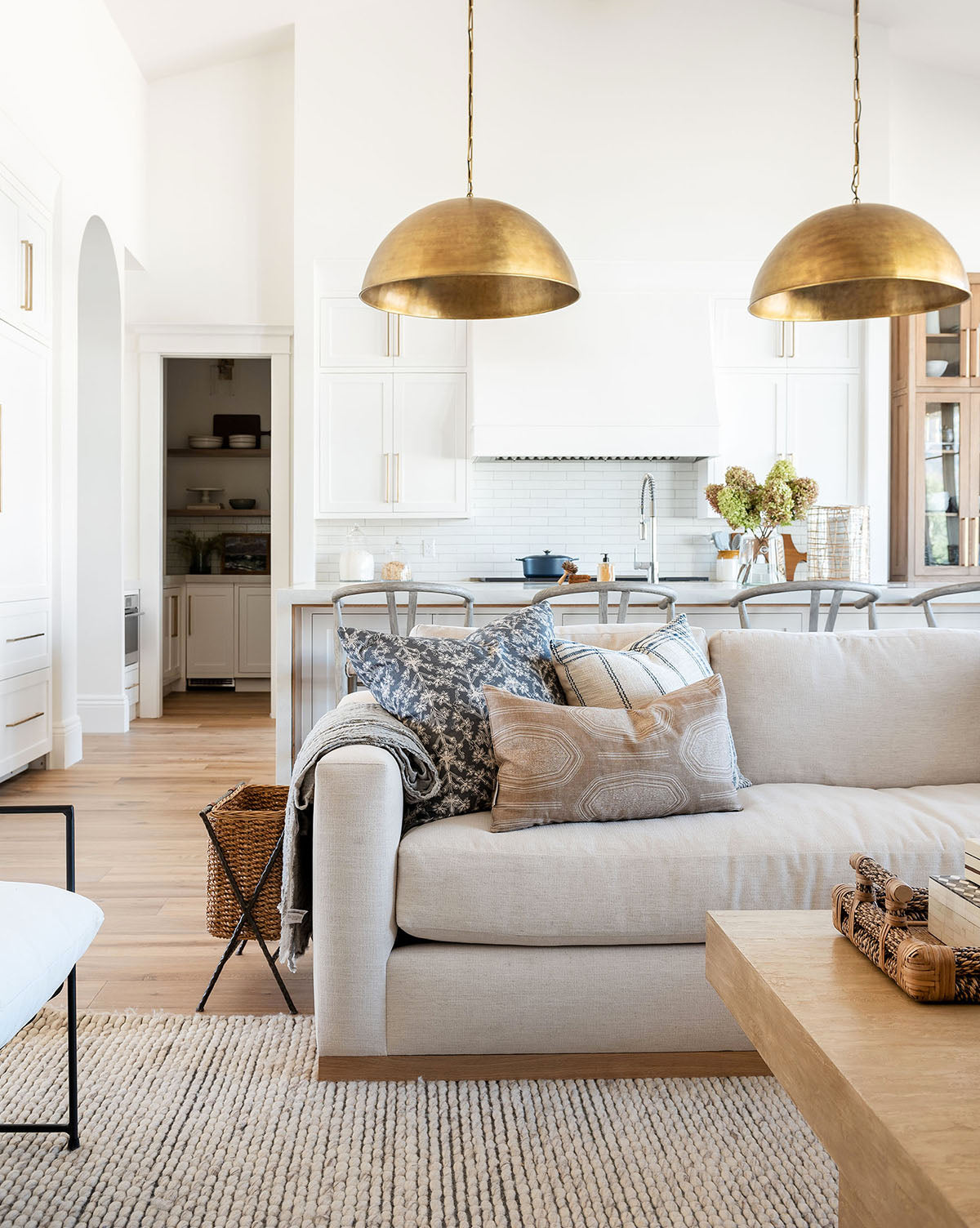 A bright, modern living room showcases a Vernon Sofa 92 with patterned pillows, a light wood coffee table, and a kitchen behind. Two large brass pendant lights hang above a white island with bar stools, by McGee & Co.