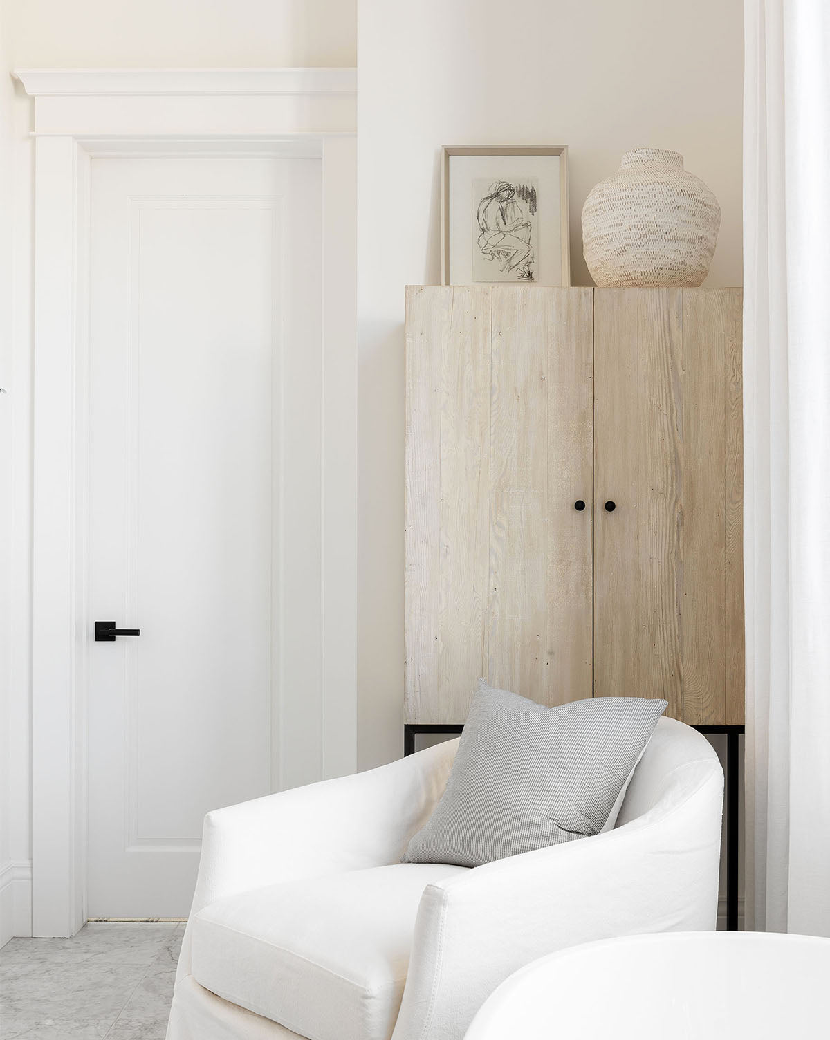 A minimalist living space featuring a white armchair with a gray pillow, light wood cabinet, ceramic vase, and the Figure Sketch framed art. The room has white walls, a door, and is filled with soft natural light.