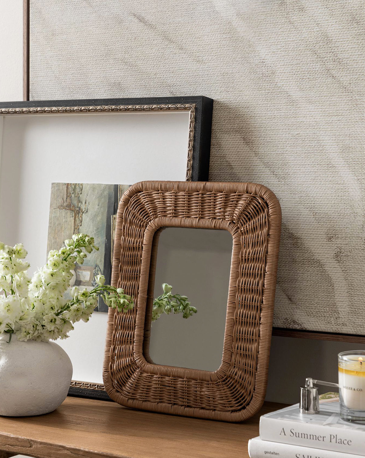 The Jera Shelf Mirror, a rectangular woven rattan piece, rests on a wooden surface beside a white flower vase, framed art print, candle, and book. Behind them hangs a large light-toned textured artwork, by McGee & Co.