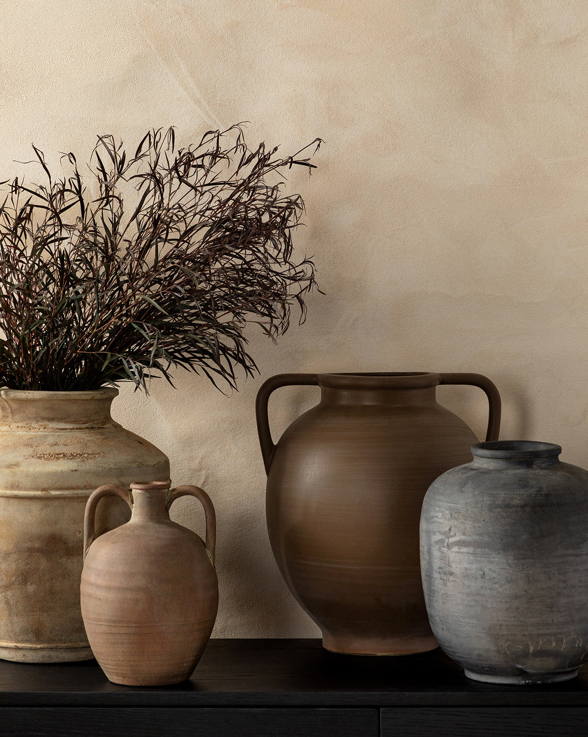 Four unique Jexa Vases in earthy tones are arranged on a dark surface against a beige wall. One robust vase holds dried branches, while the others—tan, brown, and gray—are empty and each has a distinct shape.