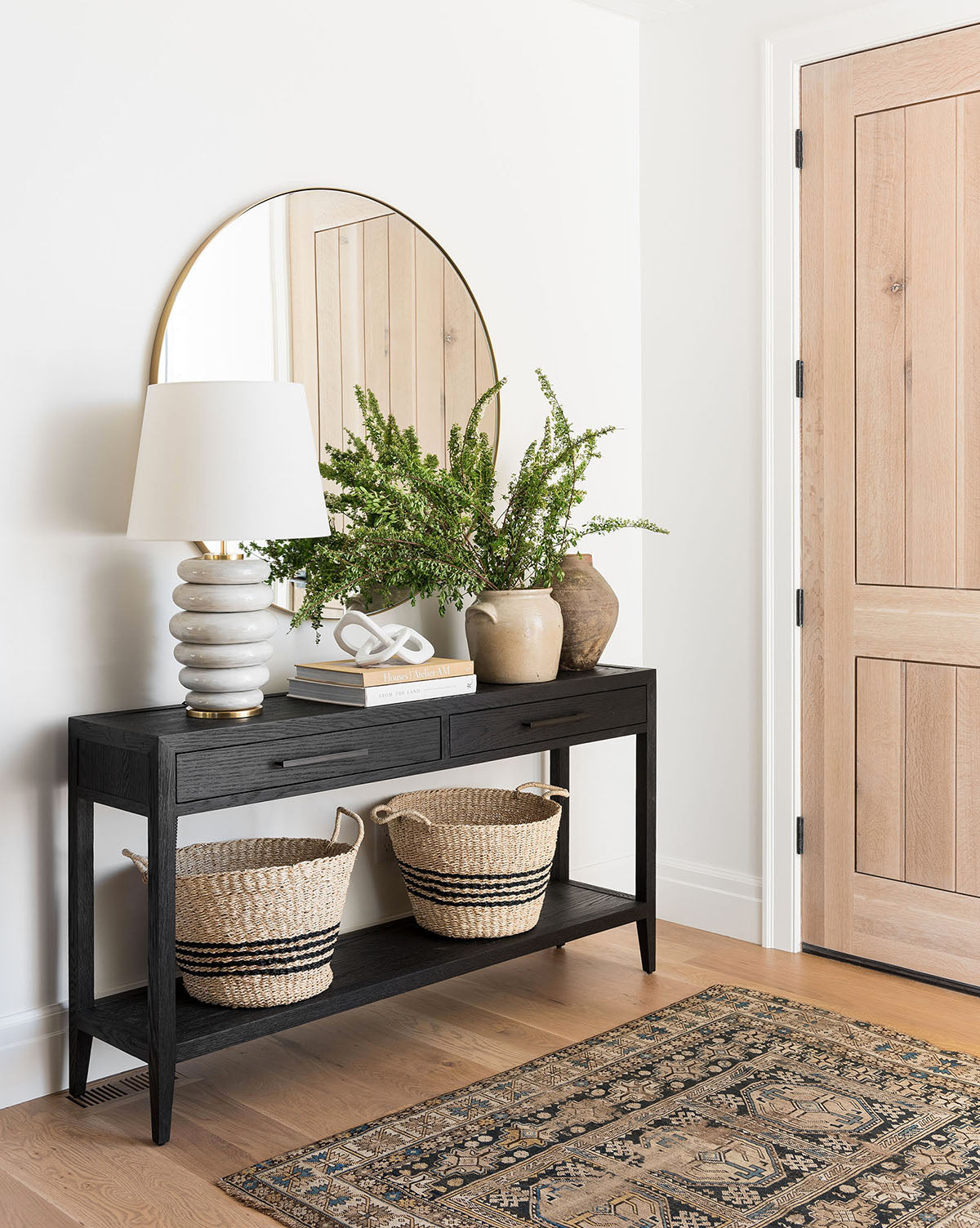 A modern entryway showcases a black console with the Phoebe Stacked Table Lamp in antiqued white, a vase of greenery, and decor. Two woven baskets rest below, while a round mirror hangs above near a light wood door, by McGee & Co.