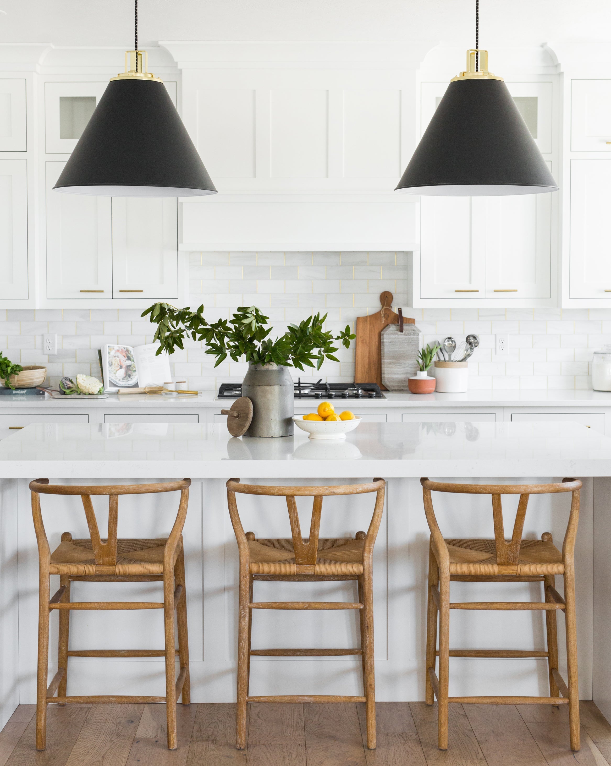 Modern kitchen with white cabinets, a large island, three Gabriel Counter Stools with rope seats and oak frames inspired by Scandinavian Modernism, black pendant lights, plus decorative touches like a bowl of lemons and green leafy branches, by McGee & Co.