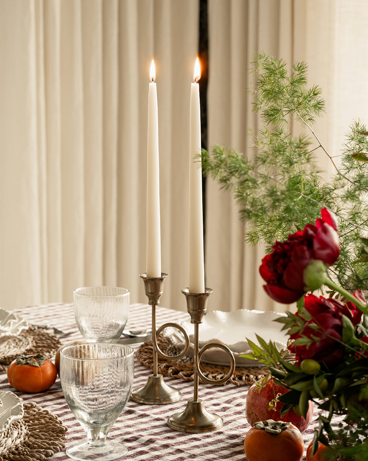 A dining table set with two lit candles in French Horn Taper Holders, clear glasses, woven placemats, red flowers, greenery, persimmons, and a pomegranate on a checkered tablecloth—ideal for elegant holiday decor, by McGee & Co.