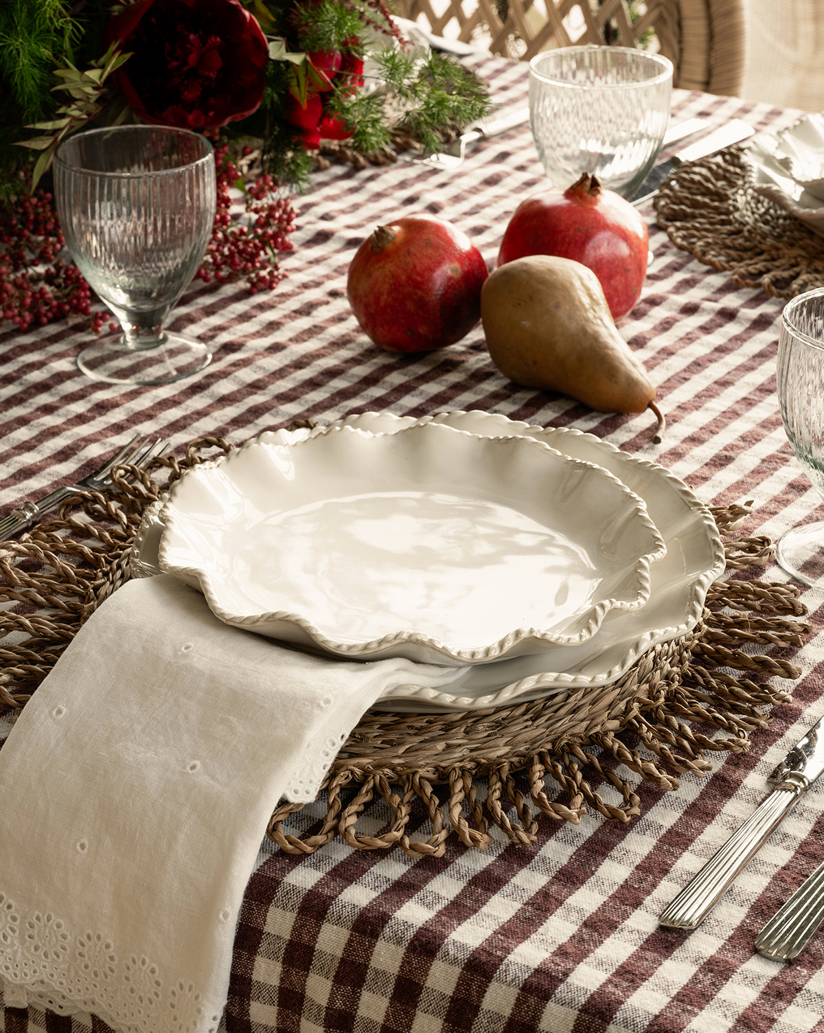 A rustic table with white ceramic plates on woven chargers, a checkered cloth, glassware, silverware, two pomegranates, a pear, greenery with red berries, and McGee & Co. x Loeffler Randall Delilah Broderie Napkins by McGee & Co.
