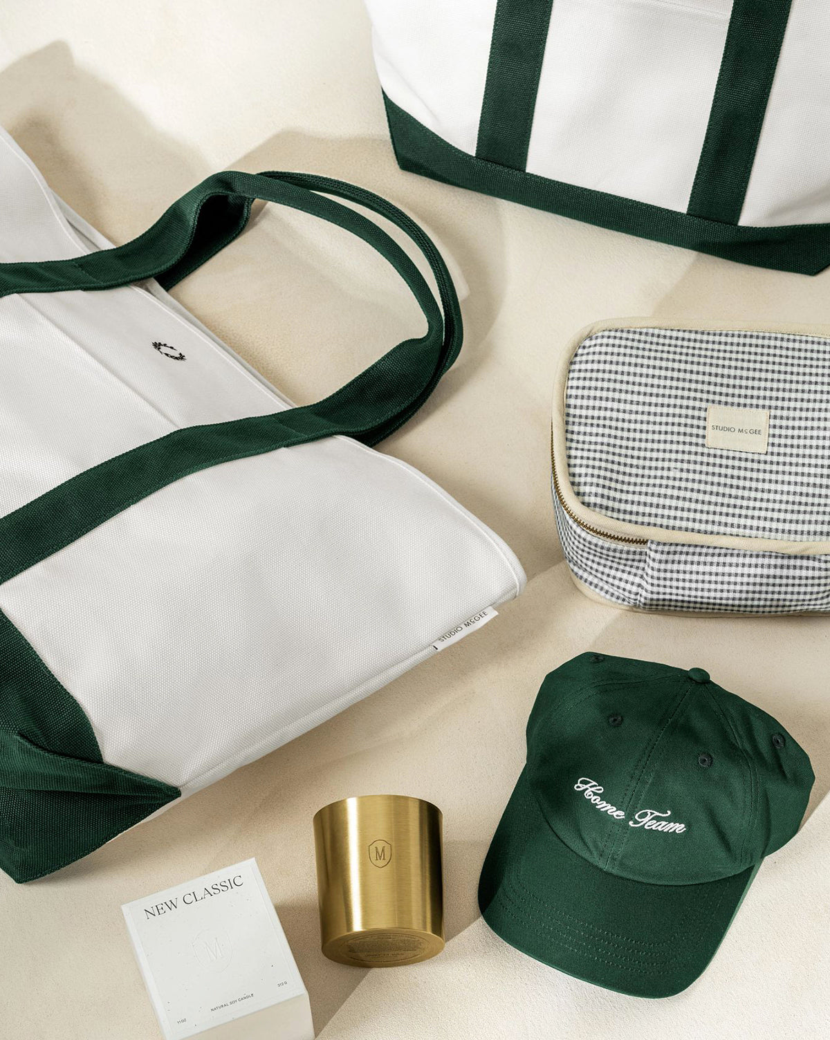 A flat lay on a beige surface showcases stylish items, including the white and green Canvas Laurel Tote, another everyday tote, a checkered pouch, a green Comme Lavie cap, a gold candle, and a white New Classic perfume box, by McGee & Co.