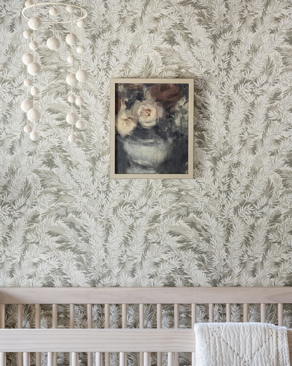 A light wood crib sits against a leafy wallpapered wall. Above, Midnight Bouquet by Leslie Dayas in a frame adds elegance, while a white felt ball mobile hangs to the left.