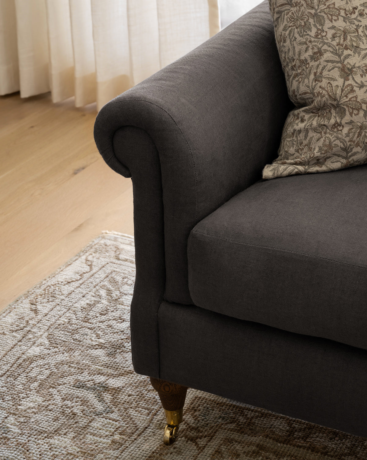 The Hildegard Sofa 90 is a dark gray upholstered, handcrafted sofa with rolled armrests, patterned cushions, and brass-capped wooden legs. It’s placed on a beige patterned rug by light curtains and a hardwood floor, by McGee & Co.