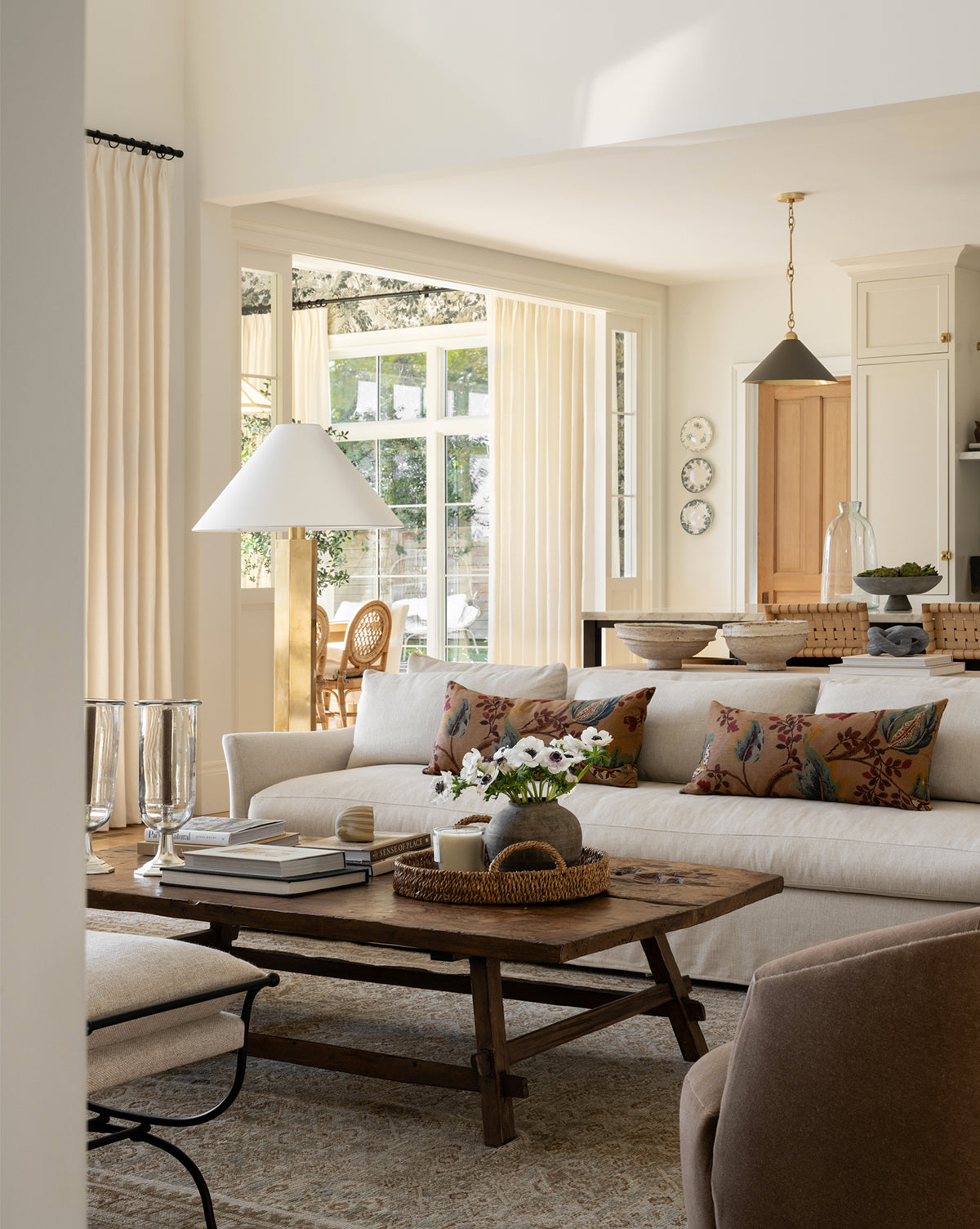 A cozy, sunlit living room features the Wilhelmina Classic Slipcover Sofa (85-110) in beige with floral pillows, a wooden coffee table topped with books and flowers, cream-curtained windows, and a dining area in the background by McGee & Co.